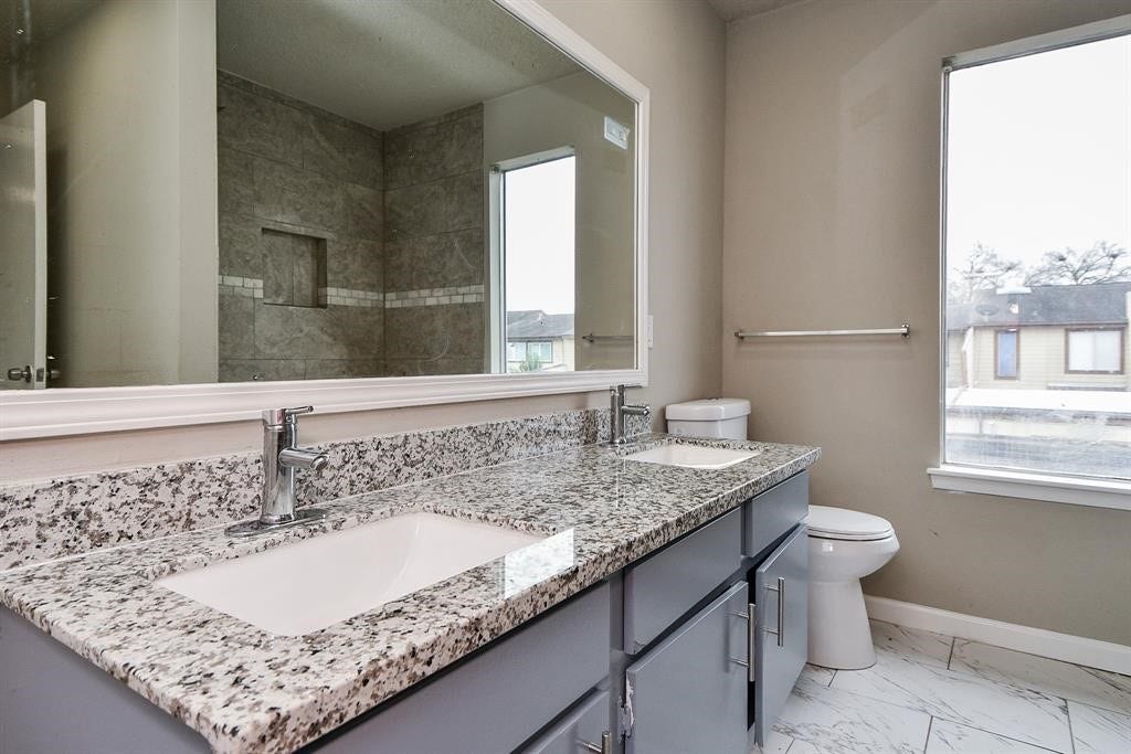 12569 Wellington Park Drive, Houston, TX 77072 This bathroom features a sleek double-sink vanity with granite countertops, modern fixtures, and a large mirror. The room is brightened by a big window and includes a tiled shower area and a toilet.