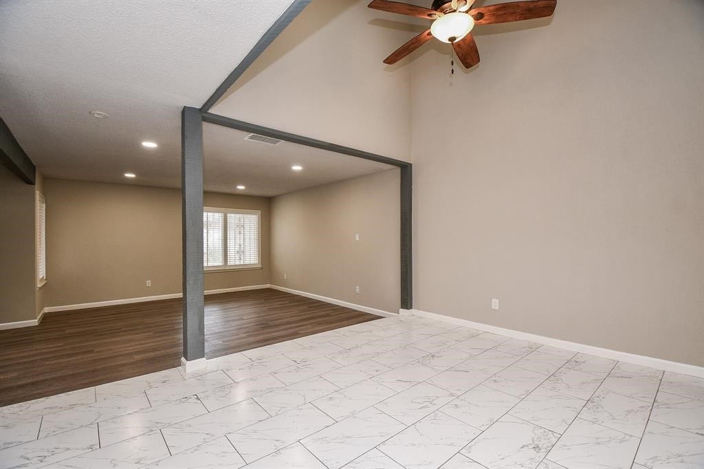 12569 Wellington Park Drive, Houston, TX 77072 This spacious room features a vaulted ceiling with a ceiling fan, a mix of wood and tile flooring, and neutral walls. The area is divided by a beam, offering distinct spaces with ample natural light from a large window.