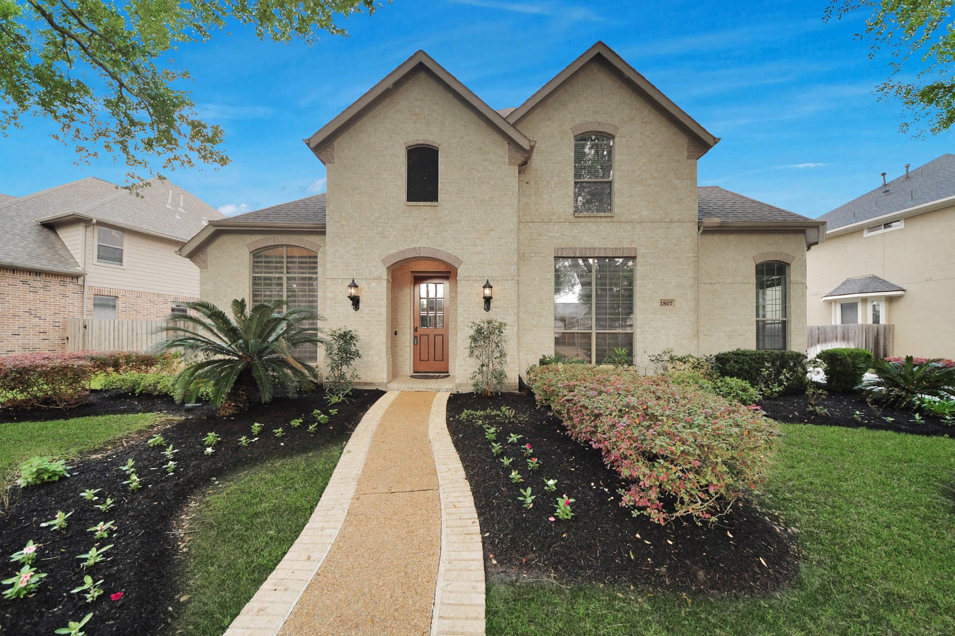 Charming two-story brick home with a welcoming front entrance. The landscaped pathway is flanked by lush greenery and vibrant plants, offering great curb appeal. Perfect for families seeking a cozy yet elegant residence.