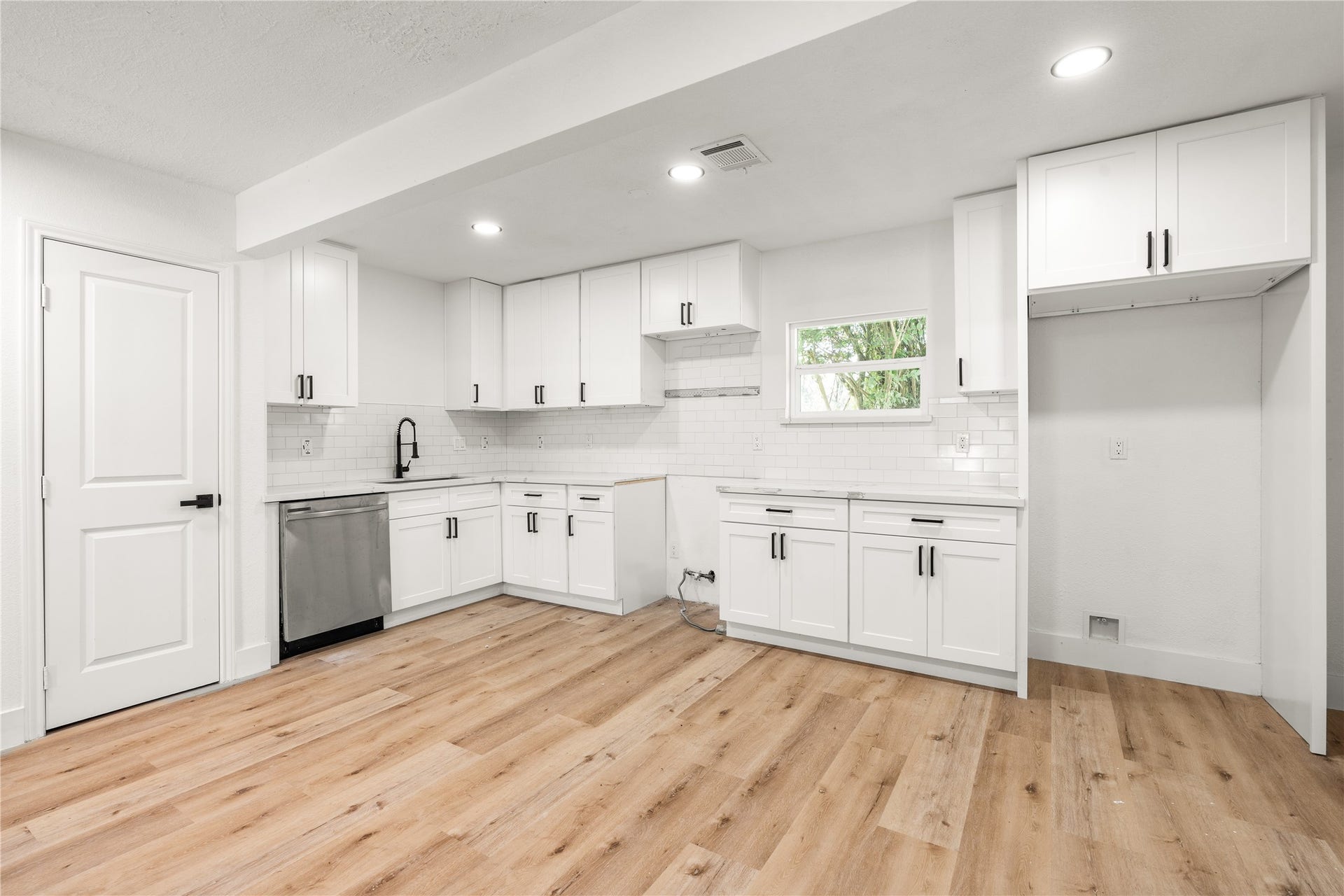 3048 Briarwick Lane, Houston, TX 77093 Full wide-angle view of the unstaged kitchen and dining area facing the main workspace, highlighting white shaker cabinets, matte black accents, subway tile, a stainless dishwasher, and continuous light wood-grain flooring.