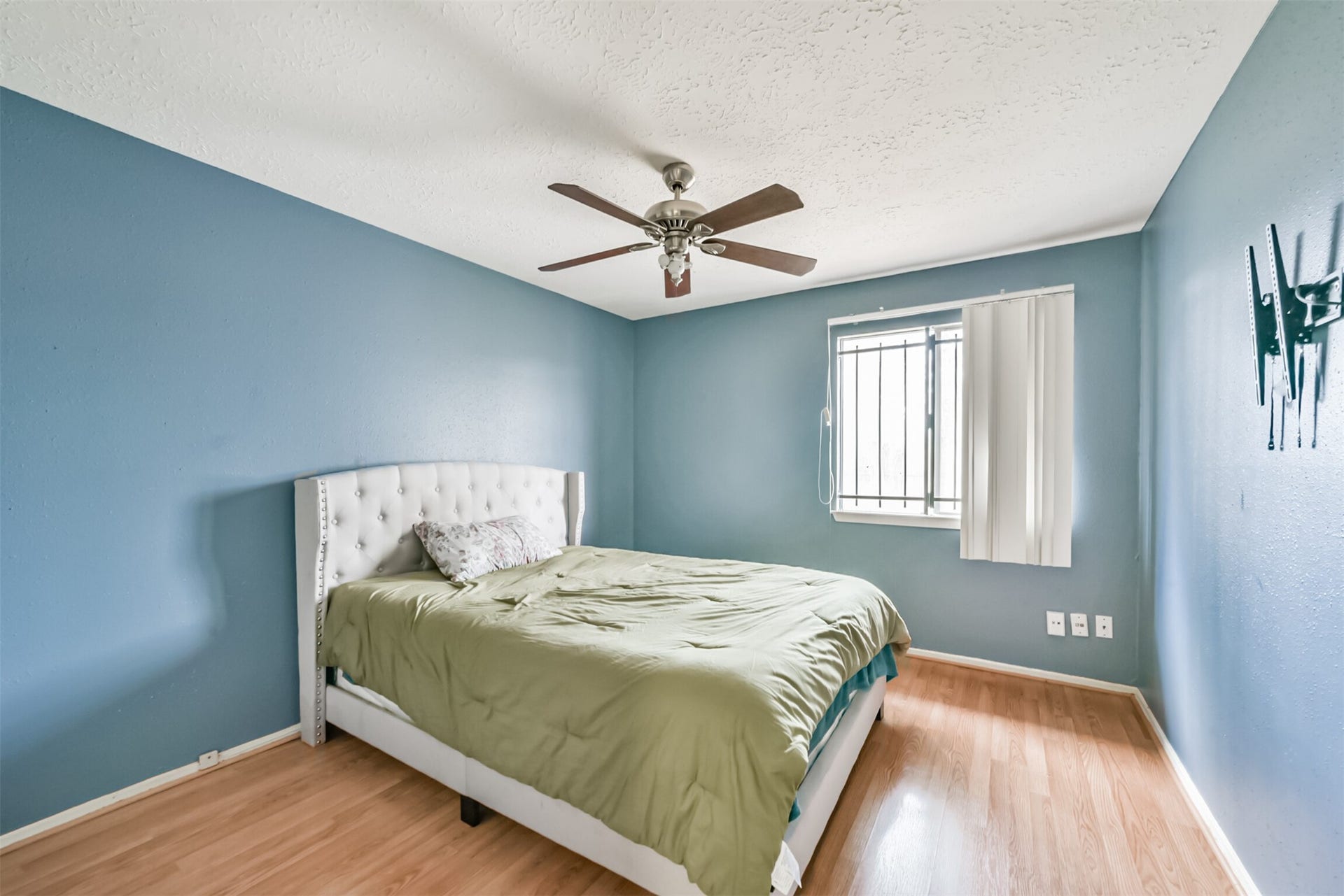 16510 Quiet Quail Drive, Houston, TX 77489 Charming bedroom with soothing blue walls, featuring a cozy bed and a ceiling fan for comfort. Natural light streams in through a window with vertical blinds, complementing the warm wood flooring.