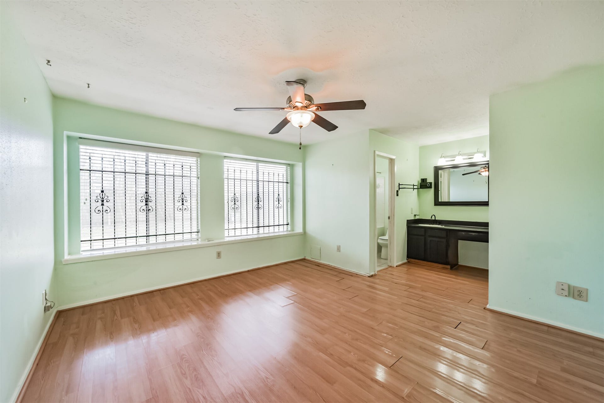 16510 Quiet Quail Drive, Houston, TX 77489 Spacious room with large windows featuring decorative ironwork, light mint green walls, and wood laminate flooring, complemented by a ceiling fan and a convenient vanity area.