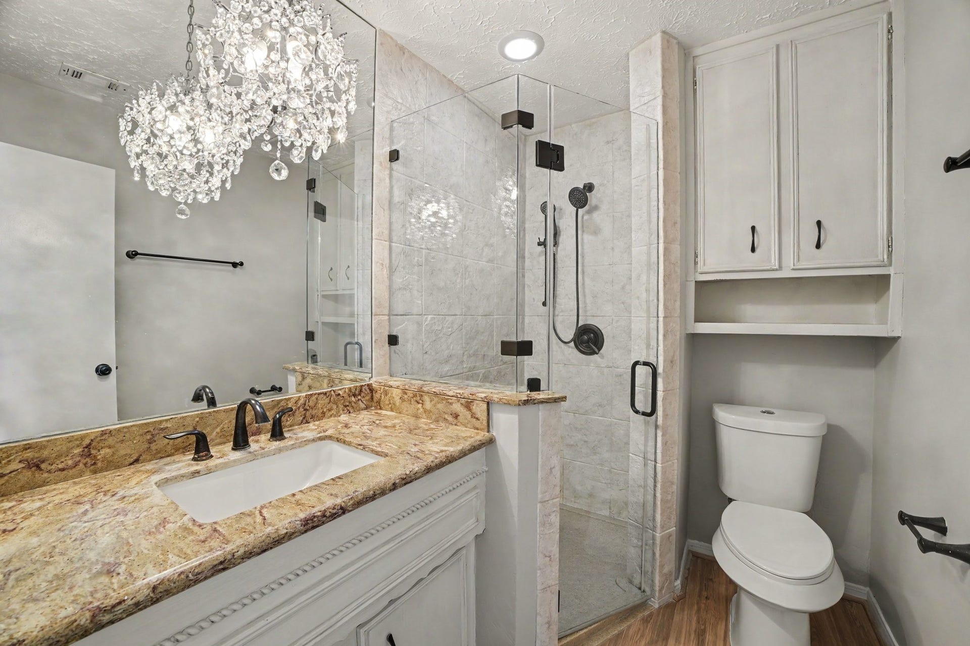 44 Chapparal Court, Missouri City, TX 77459 This luxurious ensuite primary bathroom exudes elegance with a modern frameless glass walk-in shower featuring a handheld sprayer and stylish dark matte hardware. Above the single-sink vanity, a striking crystal chandelier fixture makes a sophisticated statement, reflecting brilliantly off the large wall mirror.