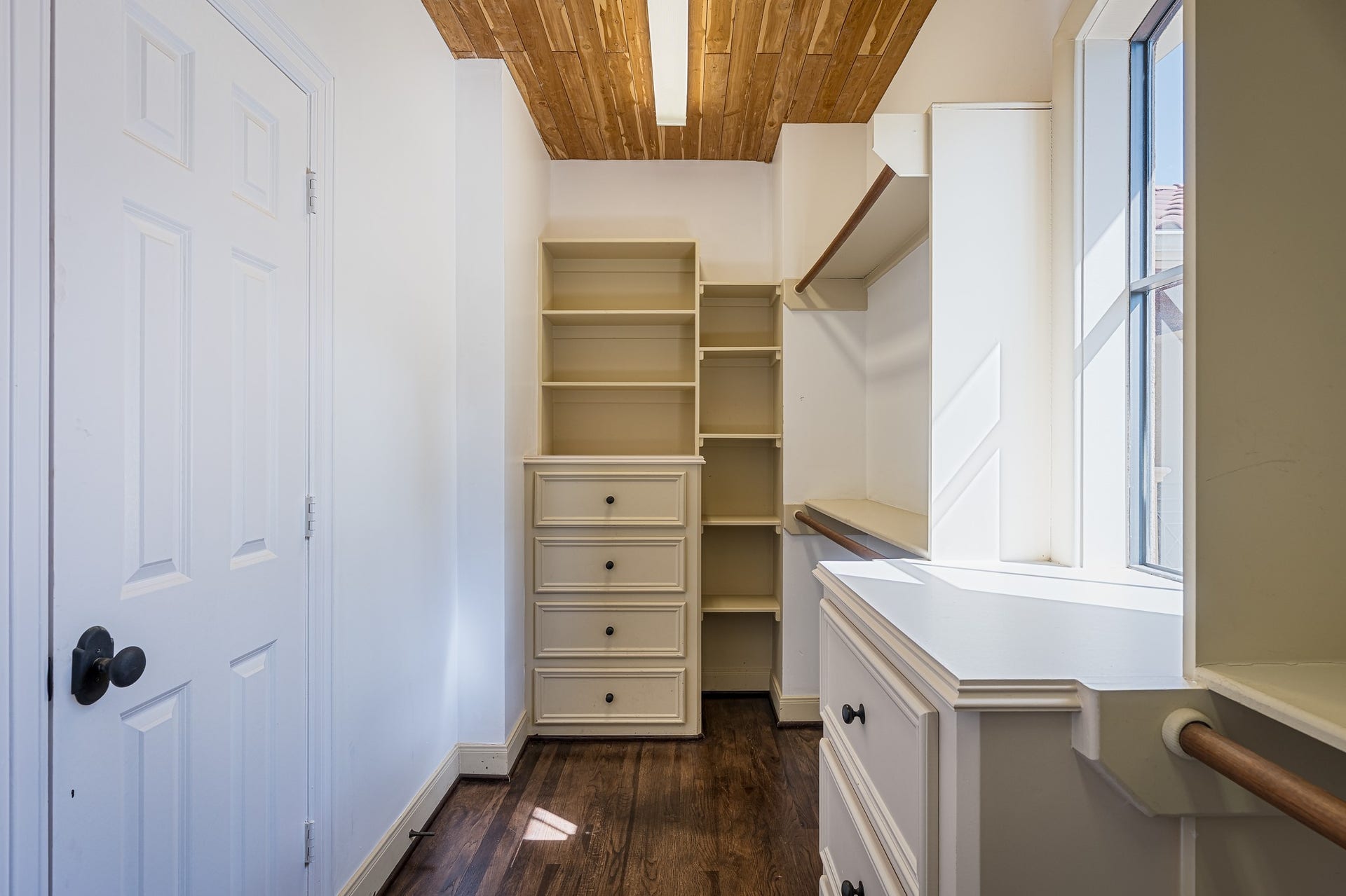 First of the two spacious walk-in primary closets with built-in shelving, drawers, and ample natural light from a large window, complemented by elegant wood flooring and ceiling.