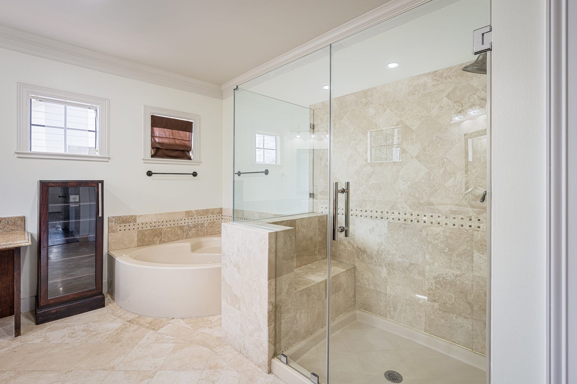 Elegant bathroom featuring a spacious glass-enclosed shower with travertine tile, a luxurious soaking tub, and ample natural light from the windows.