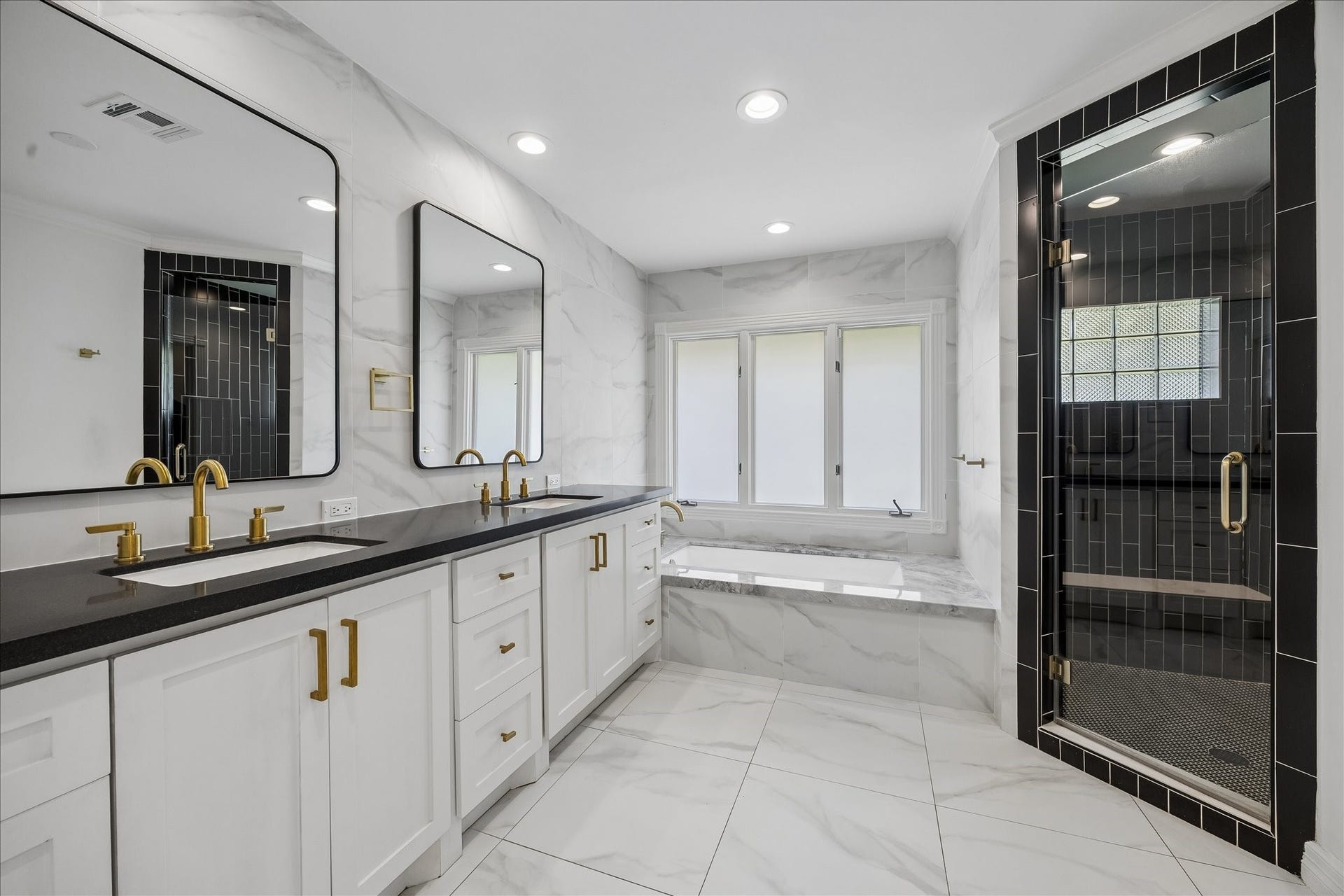 Striking primary bath features dual vanities with brass fixtures, a soaking tub and black subway tile shower, complemented by beautiful marble finishes throughout.