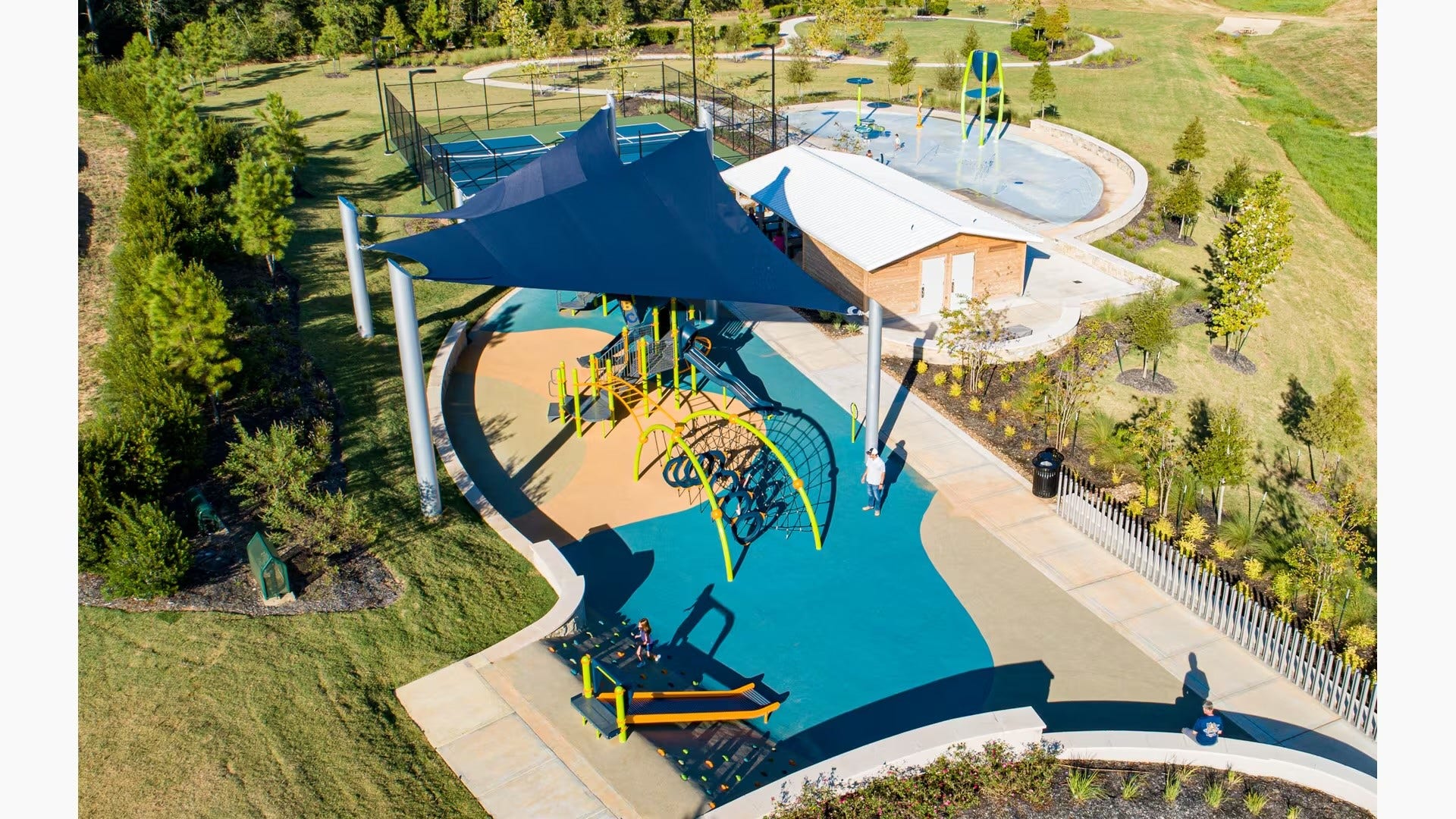 Experience vibrant outdoor living with this community park featuring a modern playground, shaded seating, tennis courts, and a splash pad, all surrounded by lush greenery.