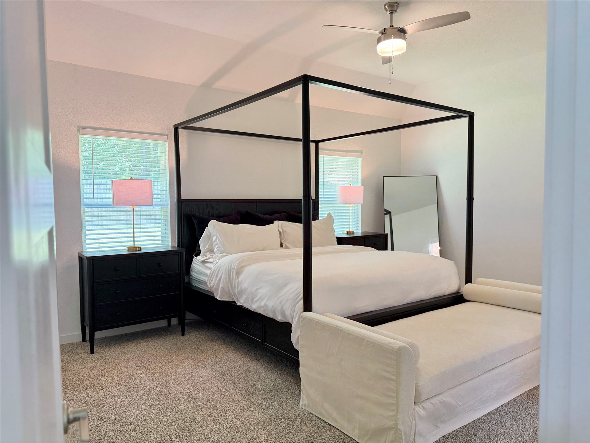 Spacious primary bedroom featuring a stylish canopy bed, plush carpeting, and ample natural light from two windows, complemented with an updated ceiling fan for comfort