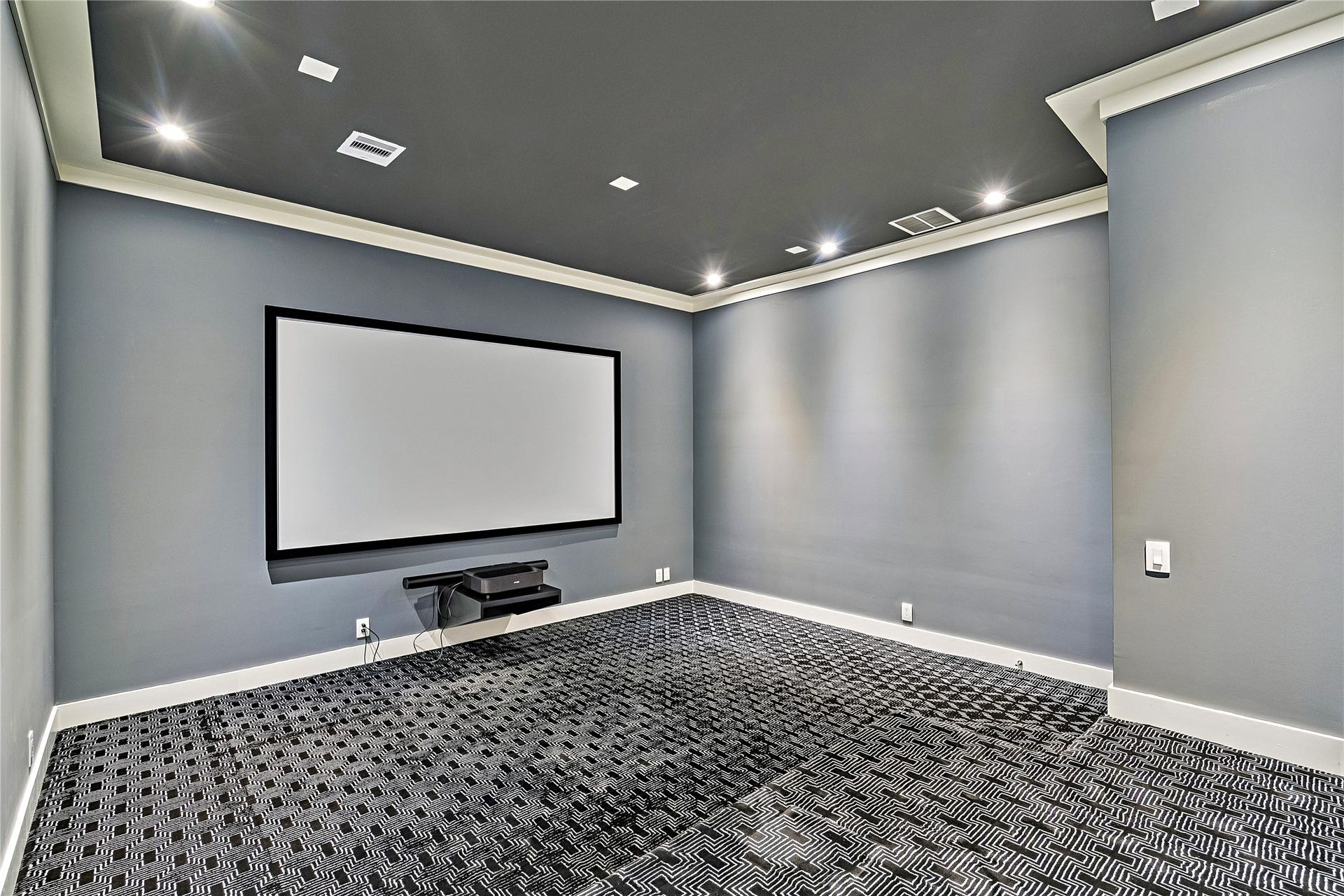 A separate media room featuring a large projection screen, dark ceiling treatment, recessed lighting, and patterned carpeting. Designed for private movie screenings and entertainment away from the home's principal living areas.