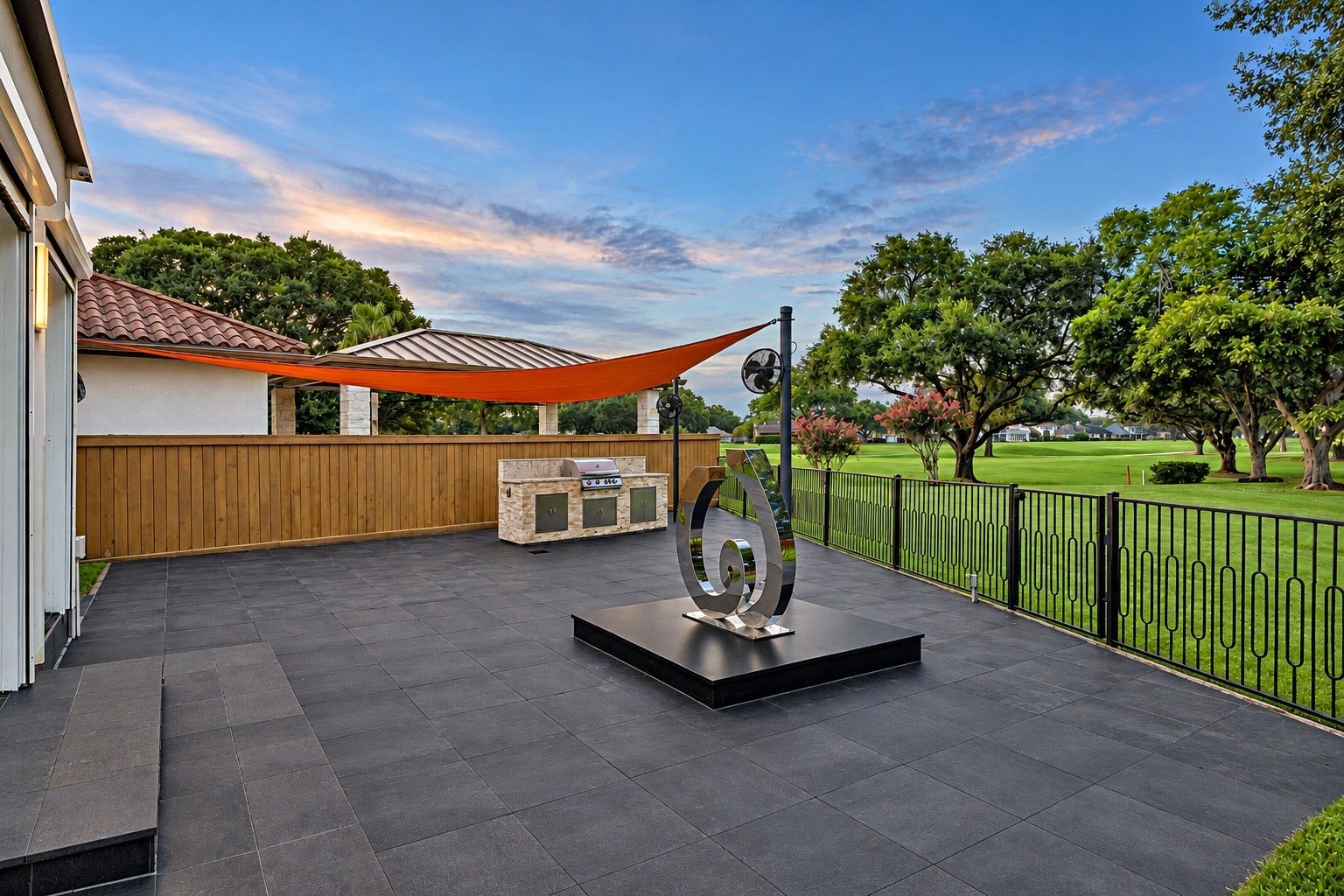 An exceptional outdoor entertaining area featuring expansive hardscaping, an outdoor cooking station, architectural shade sails, and a polished-steel sculpture by an Argentine artist. The terrace overlooks mature landscaping and the fairways of Sugar Creek Country Club, creating a distinctive setting for outdoor gatherings.