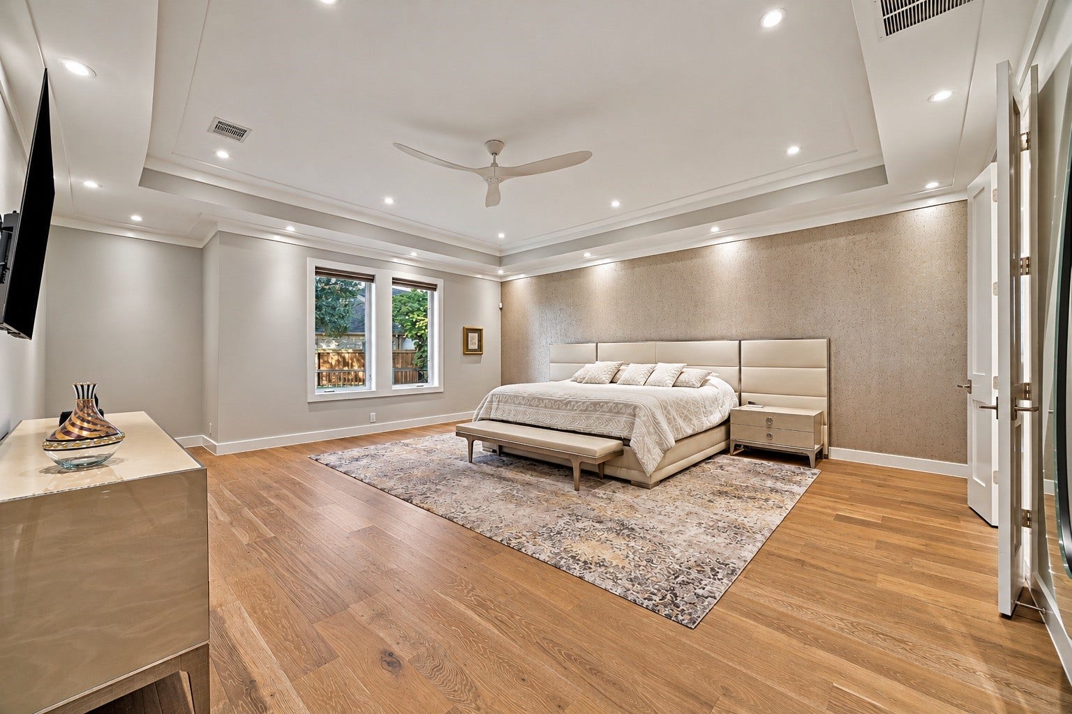 An expansive primary bedroom featuring beautiful wood flooring, recessed lighting, and a sophisticated neutral palette. The first-floor location provides a private retreat separate from the home's upper-level bedrooms and recreational spaces.