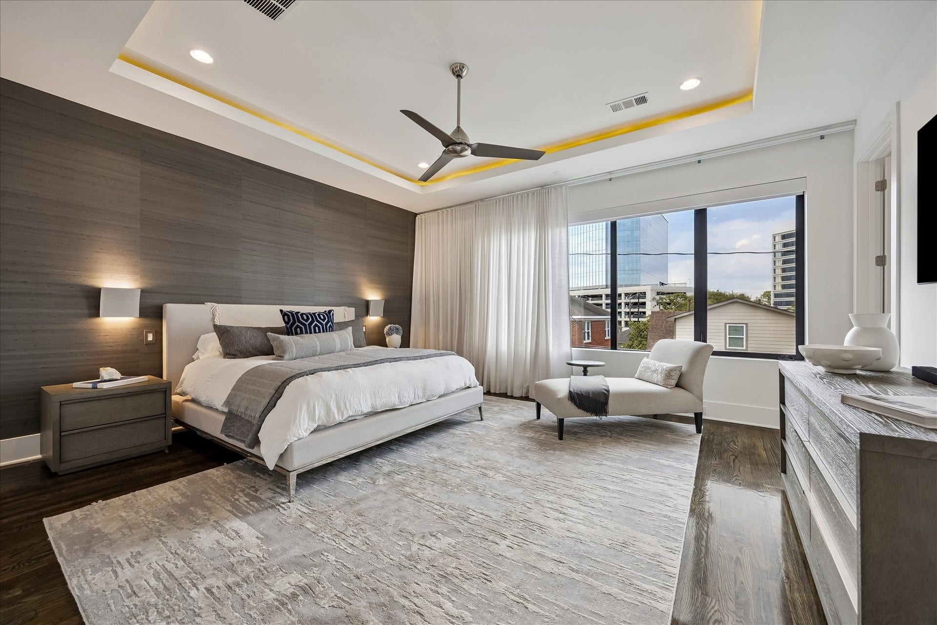 Luxurious primary bedroom with an elegant coved ceiling and silk wallpaper accent wall. Enjoy linen drapes and Lutron-controlled blackout shades for perfect light and privacy. Features a spacious closet with floor-to-ceiling storage and a 65-inch Samsung TV for ultimate style and convenience.