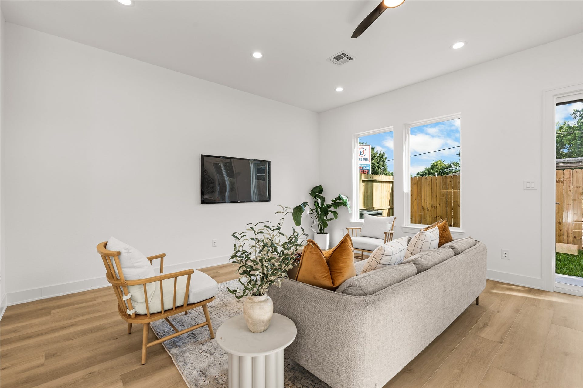 This view shows the spacious layout and natural light of the main living room. The space easily fits a sofa and wood accent chairs, while large windows look out into the private backyard. A wall-mounted TV demonstrates the room's setup for entertainment.