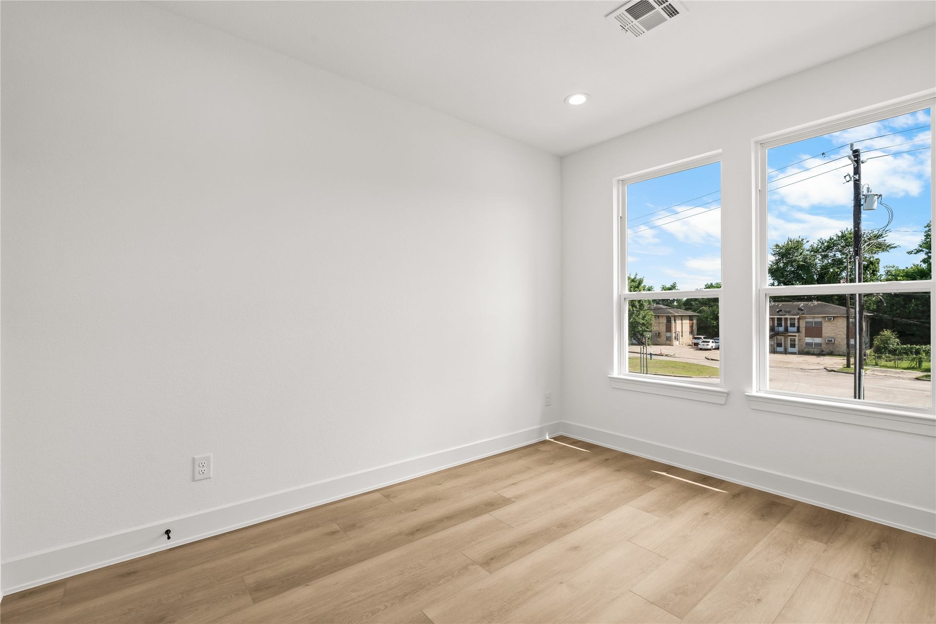 The second bedroom features a bright, airy layout with white walls and warm flooring. Two large vertical windows provide plenty of natural light and neighborhood views, while the spacious layout easily fits a queen-sized bed and extra storage furniture.