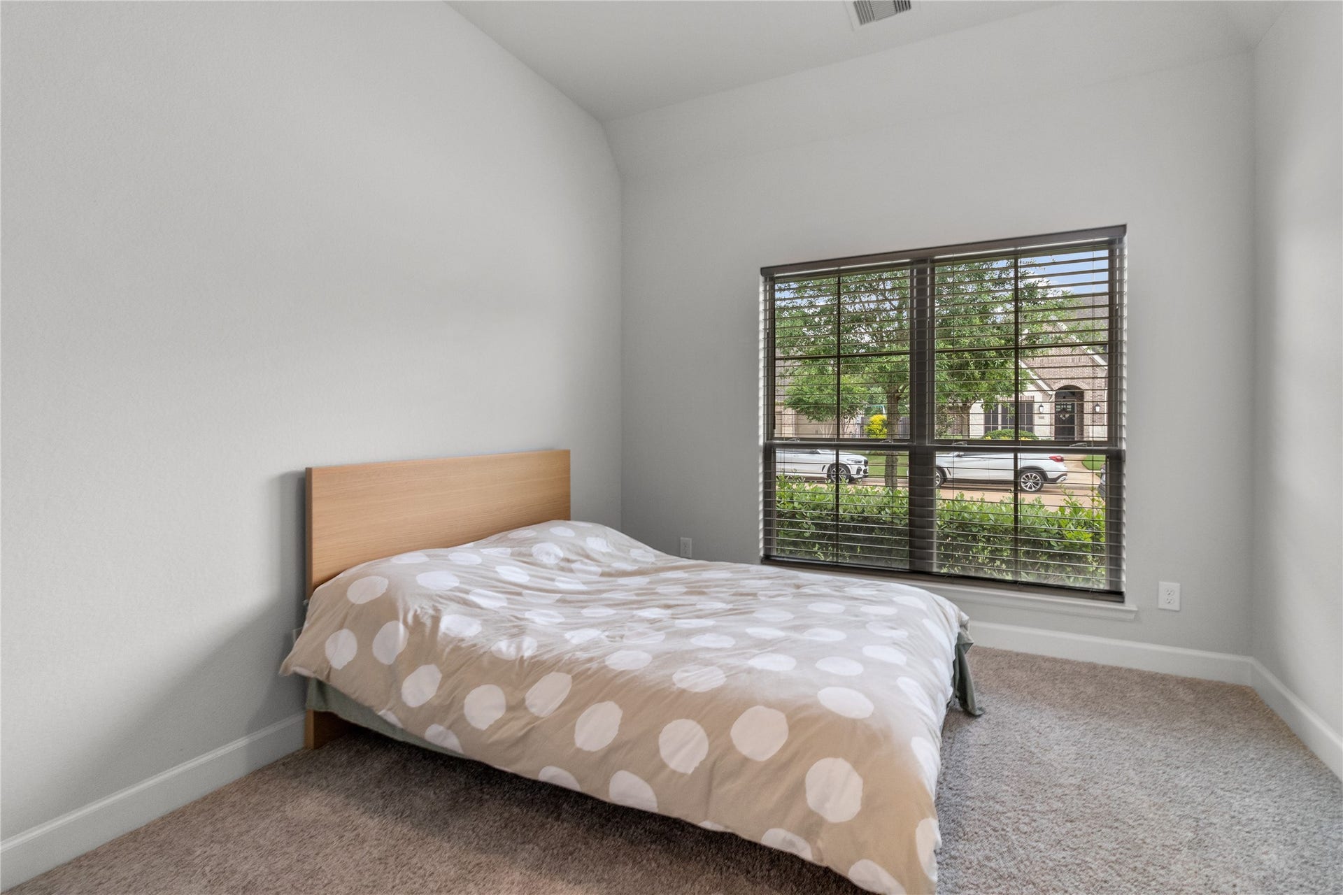 30427 Watershed Way, Fulshear, TX 77441 Cozy bedroom with a neutral color palette, featuring a bed with a polka dot comforter. The large window with blinds lets in natural light and offers a view of the front yard and neighborhood.