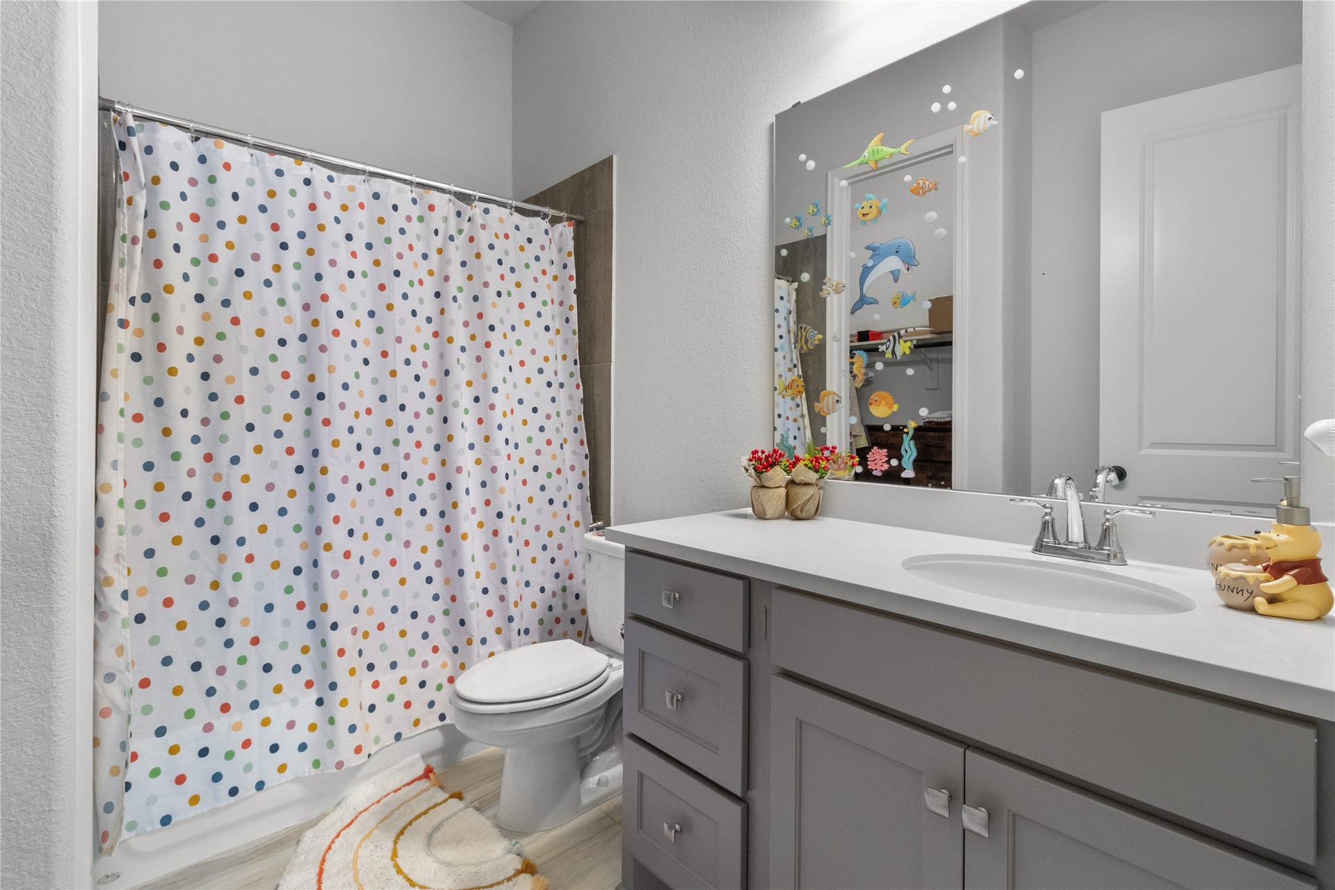 30427 Watershed Way, Fulshear, TX 77441 This bathroom is bright and cheerful, featuring a colorful polka dot shower curtain and kid-friendly wall decals. It has a modern gray vanity with a sleek white countertop and ample storage.
