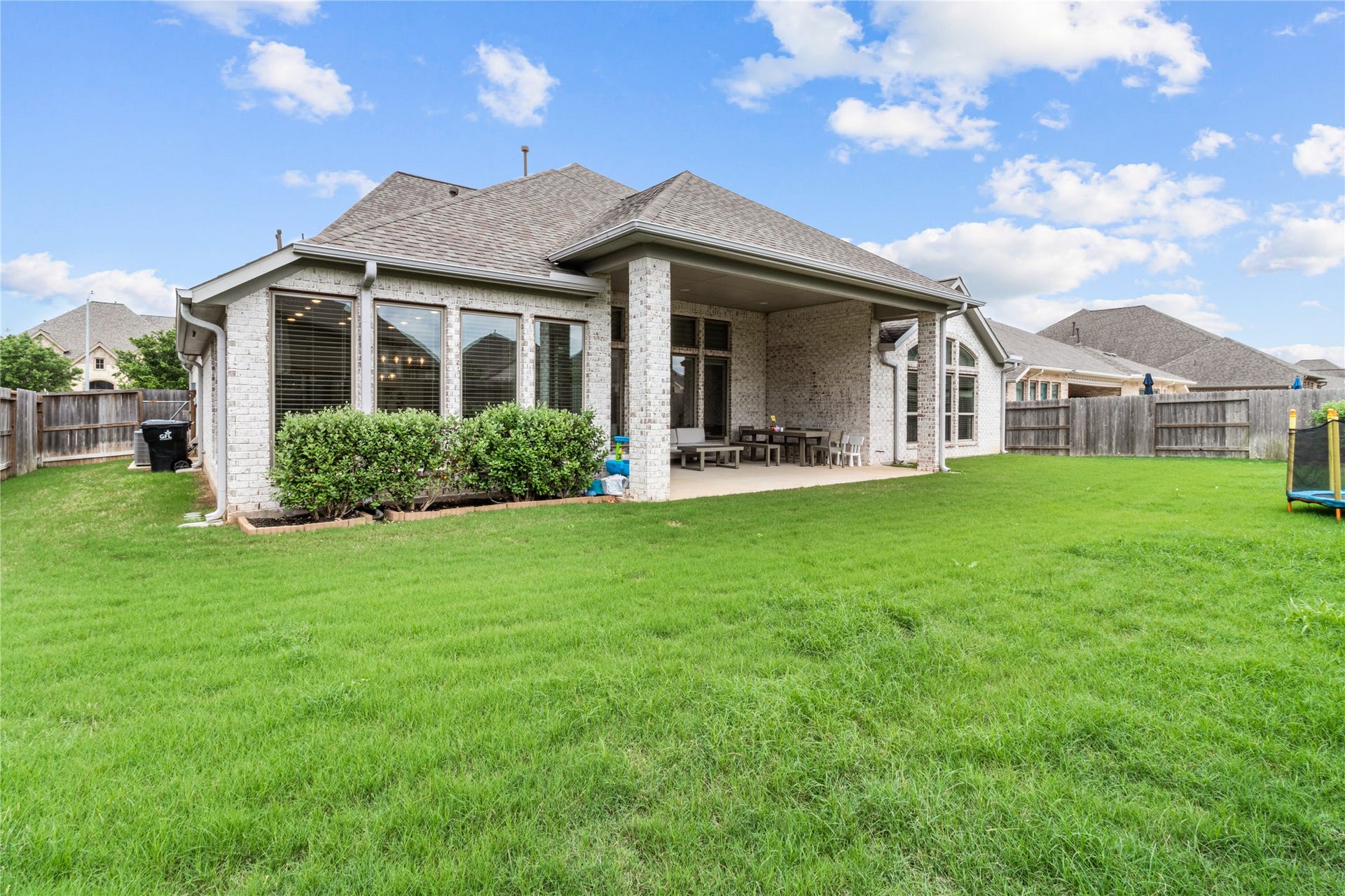 30427 Watershed Way, Fulshear, TX 77441 A spacious backyard with lush green grass, a covered patio perfect for outdoor entertaining, and large windows for ample natural light. The home exterior is a charming brick design, within a fenced area offering privacy.