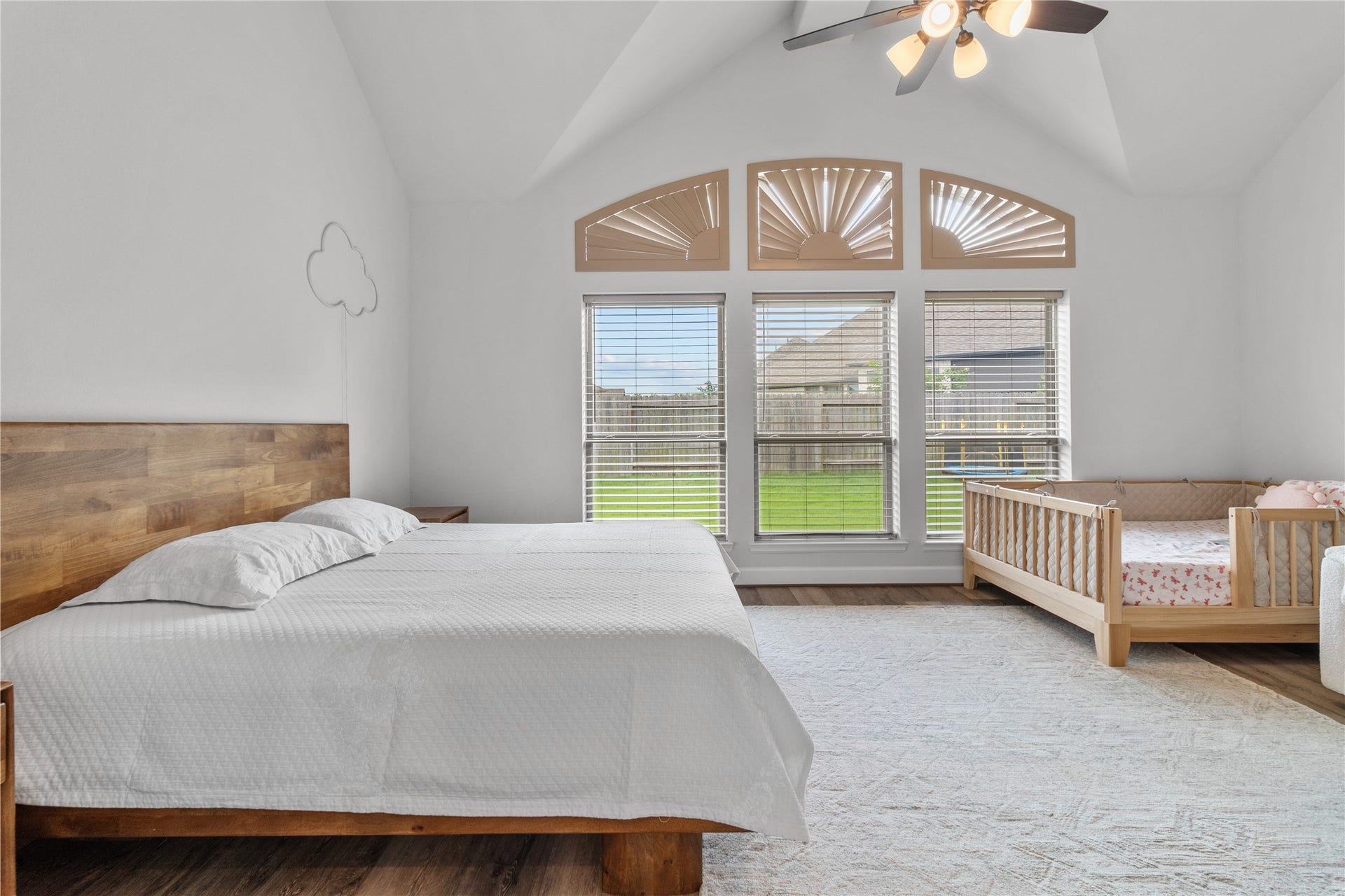 30427 Watershed Way, Fulshear, TX 77441 This spacious bedroom features high ceilings and large arched windows that allow plenty of natural light. It includes a stylish wooden bed and a cozy crib, perfect for a family setting. The room has a warm, inviting atmosphere with a view of a green backyard.