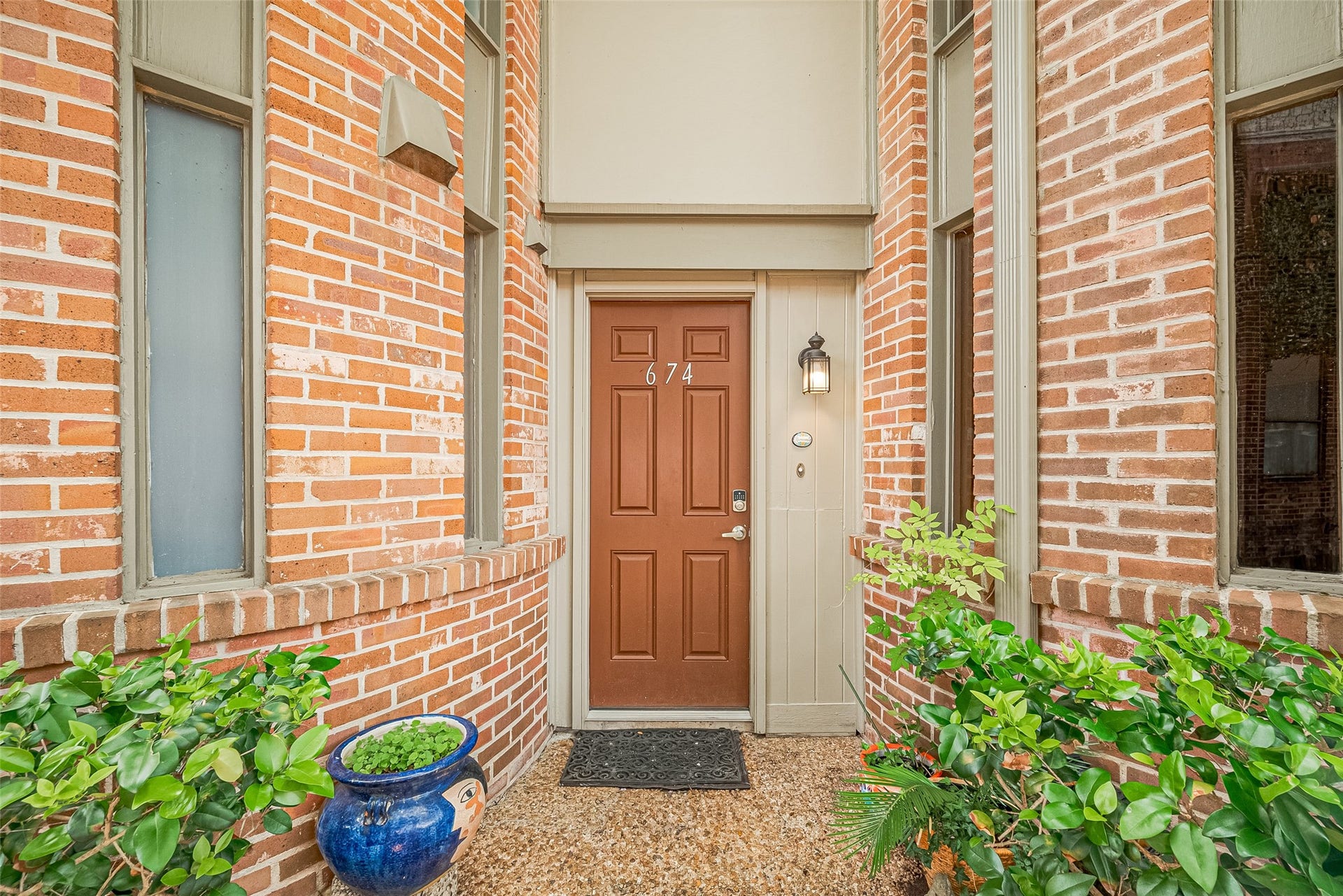 Charming brick entryway features a welcoming front door framed by large windows, complemented by lush greenery and a classic lantern light.