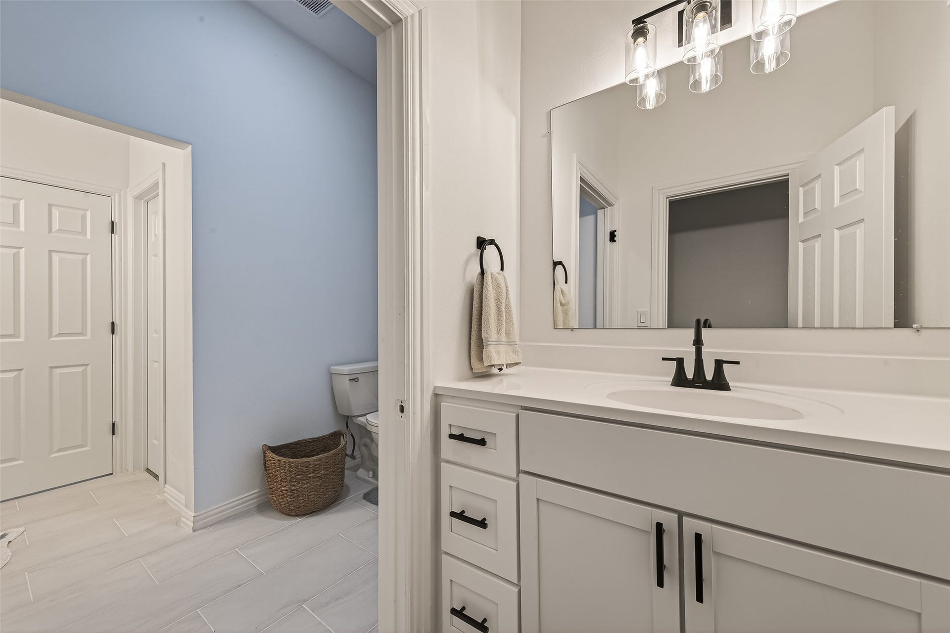 4423 Parkwater Cove Court SE, Sugar Land, TX 77479 This bathroom features a modern, clean design with a white vanity, black fixtures, and a large mirror. It includes a separate toilet area with light blue walls and tiled flooring, offering a fresh and inviting space.