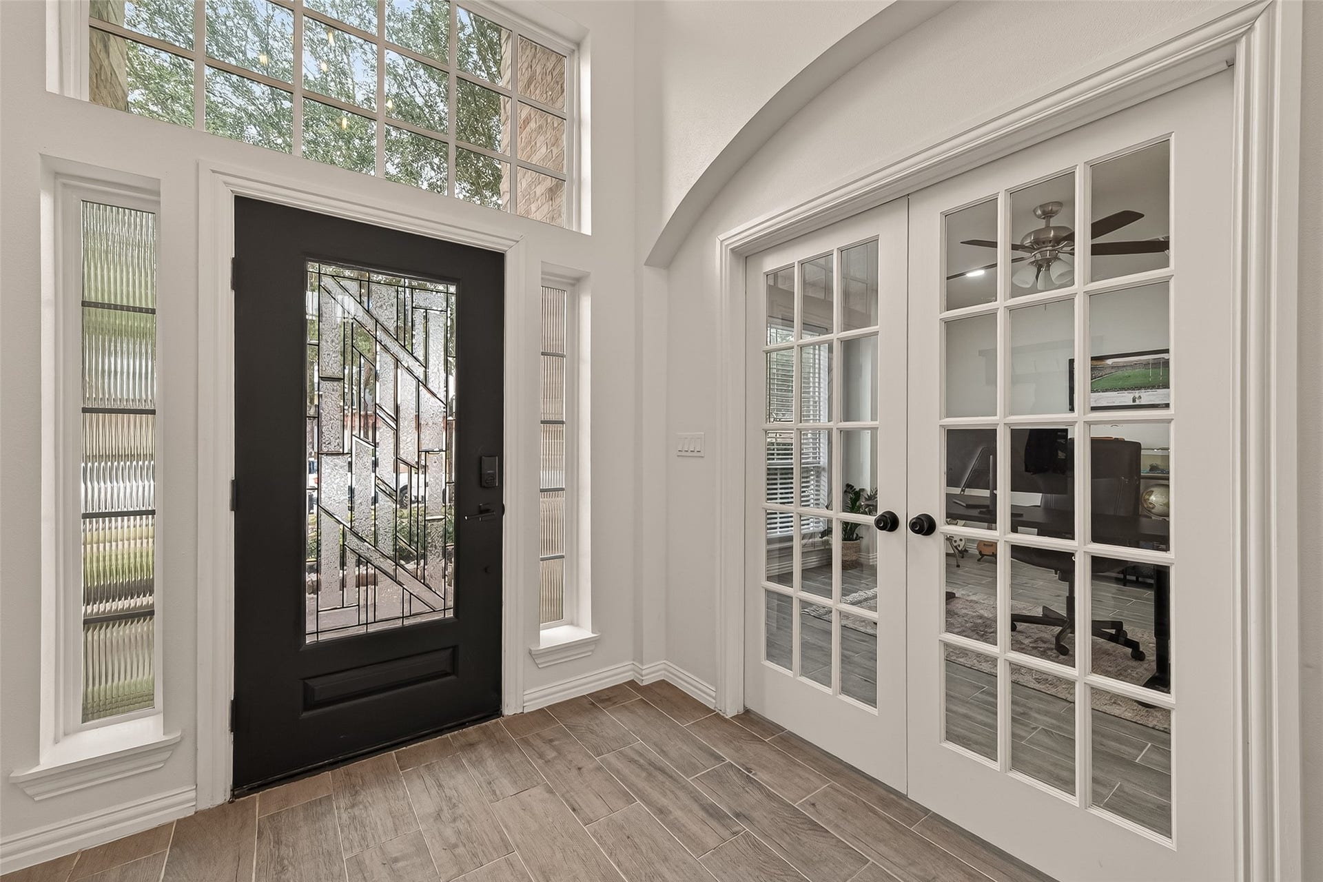 4423 Parkwater Cove Court SE, Sugar Land, TX 77479 This entryway with high ceilings features a stylish black door with decorative glass panels, flanked by tall windows for natural light. Adjacent is a set of elegant French doors leading to a bright room with a ceiling fan, ideal for an office or study. The space is enhanced by neutral walls and wood-look tile flooring.