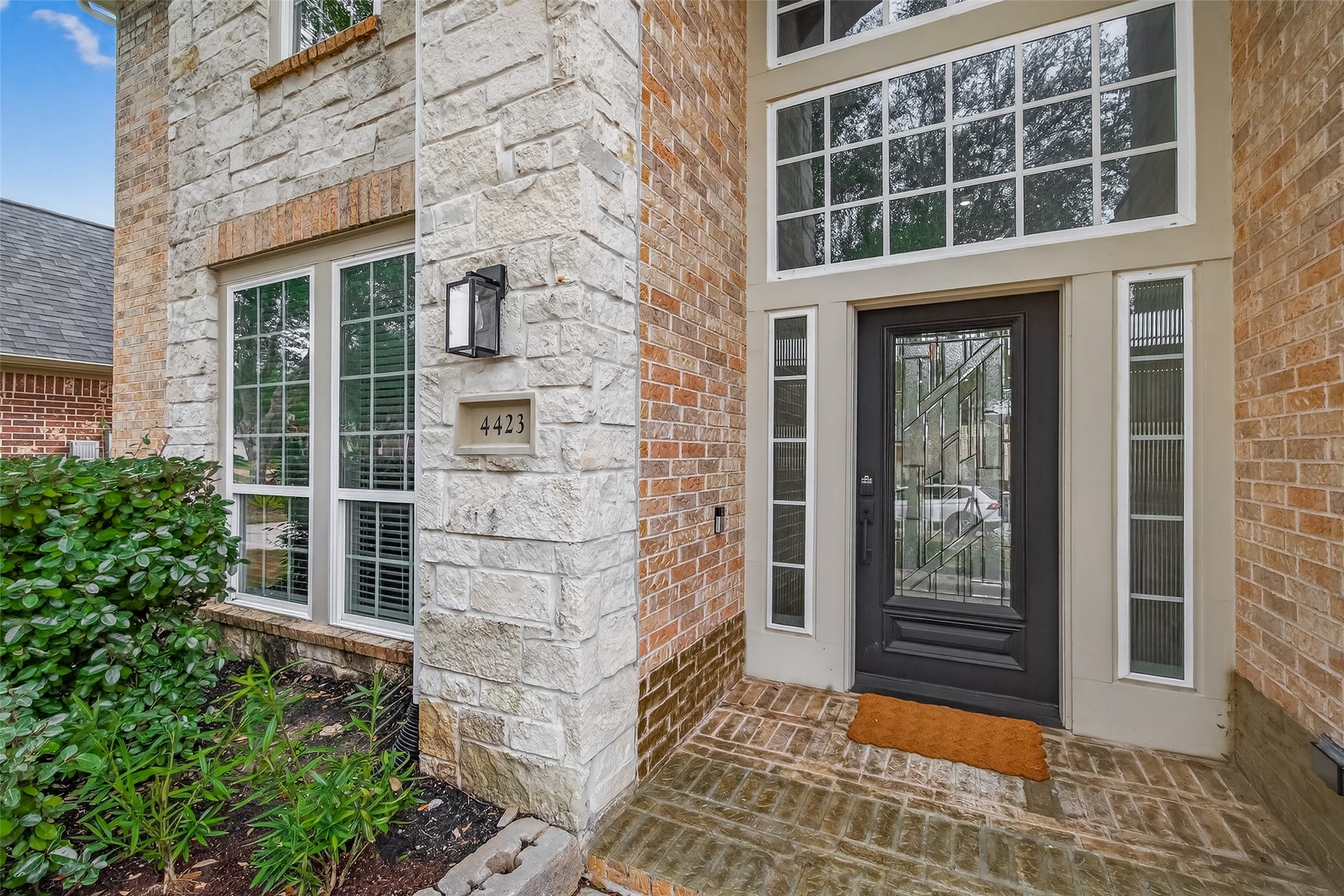 4423 Parkwater Cove Court SE, Sugar Land, TX 77479 This photo showcases the elegant entrance of a home, featuring a black door with decorative glass panels, surrounded by large windows for ample natural light. The exterior combines brick and stone, with well-maintained landscaping enhancing curb appeal.