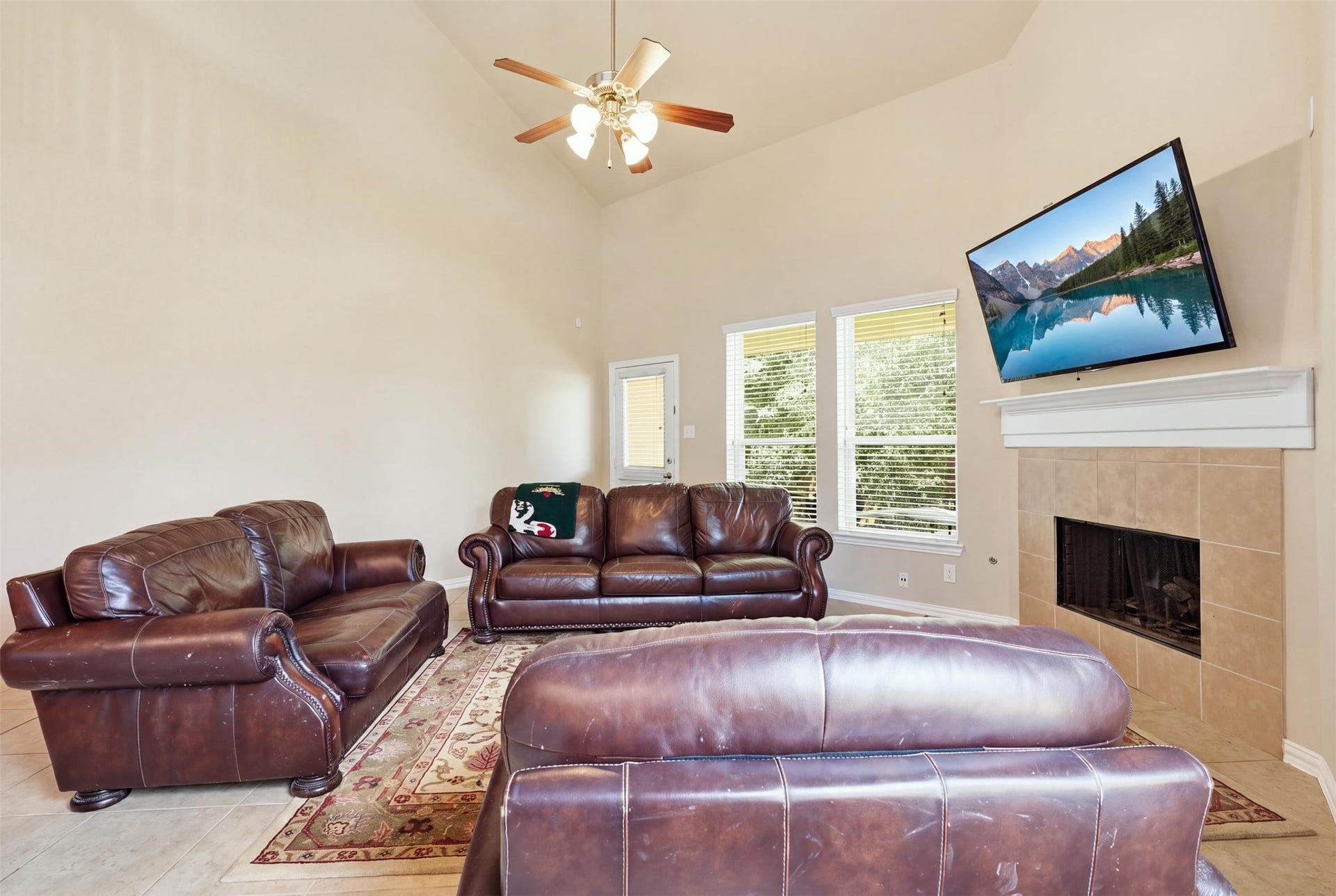 Big-sky living room: soaring ceilings, a gas-log fireplace with tile surround and mantel, and a wall of windows framing the green view out back. The door to the covered patio is right here.