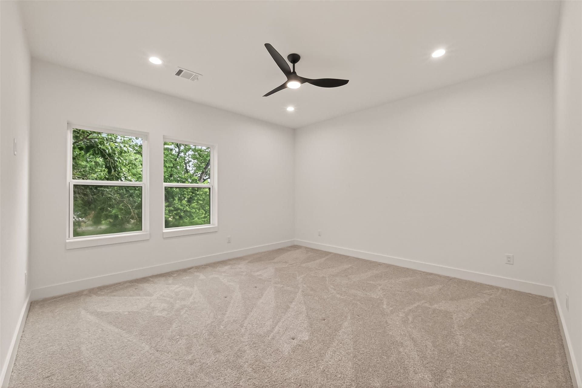 6808 Madrid Street, Houston, TX 77021 This room features clean white walls, a modern ceiling fan, and plush carpeting. Natural light streams through two large windows, offering a view of lush greenery, creating a bright and inviting space.