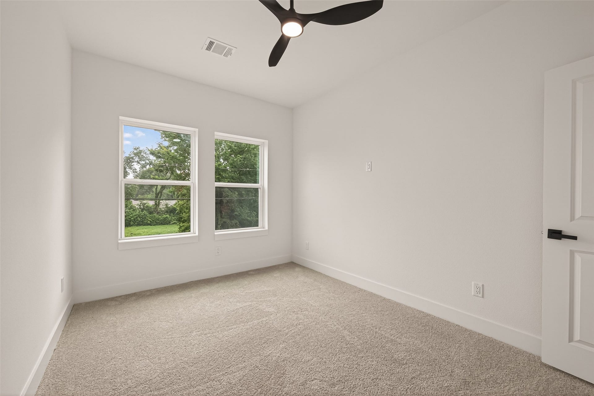 6808 Madrid Street, Houston, TX 77021 This is a bright, cozy room with two large windows offering a view of lush greenery. It features clean, neutral walls, a modern ceiling fan, and soft carpet flooring, creating a welcoming atmosphere.