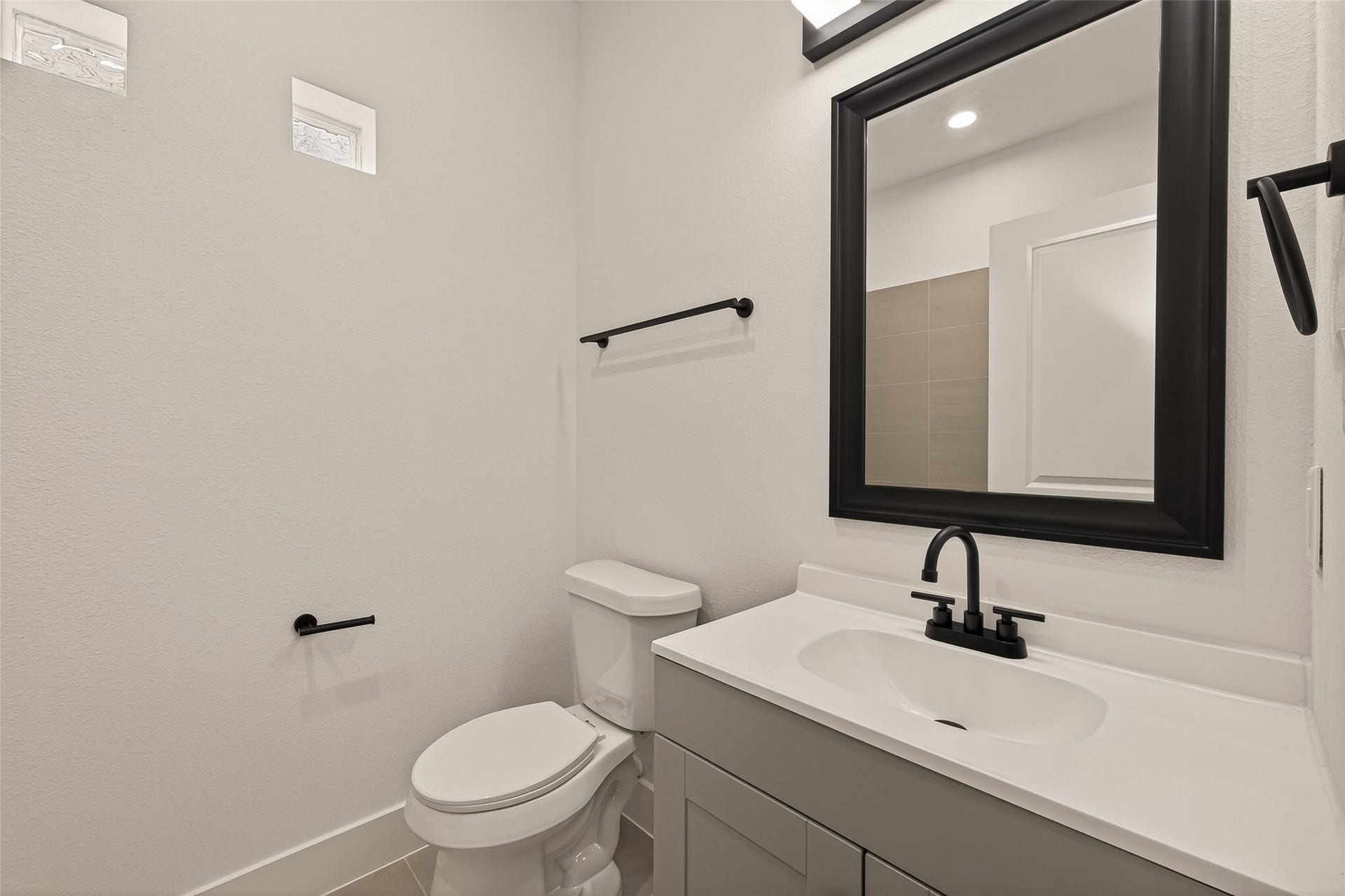 6808 Madrid Street, Houston, TX 77021 This bathroom features a clean, modern design with a white vanity and black fixtures. It includes a toilet and a large mirror above the sink, complemented by neutral tile flooring and two small windows for natural light.
