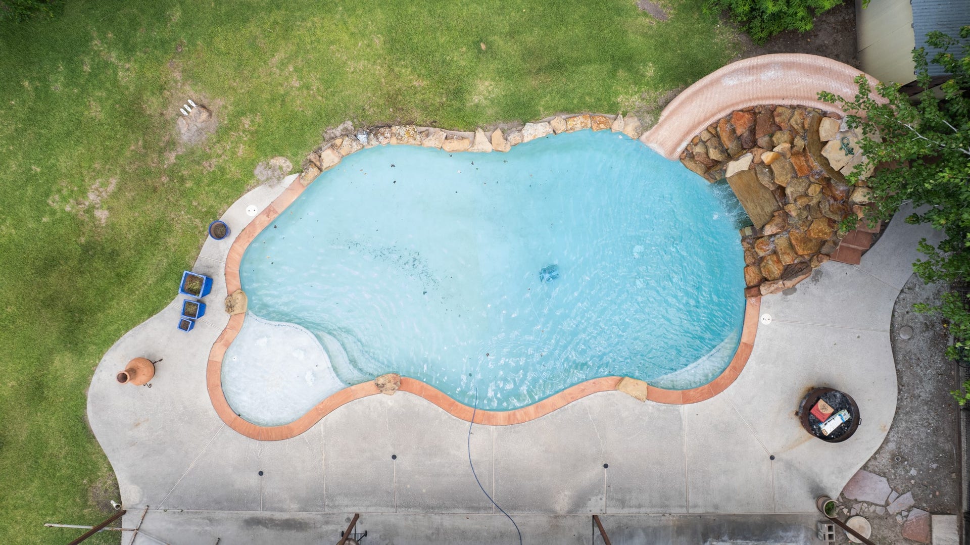 15602 Fir Road, Santa Fe, TX 77517 Stunning aerial view of the resort-style pool showcasing the custom rock waterfall slide, expansive concrete decking, and generous outdoor entertaining space. The freeform design and surrounding green space create a true backyard oasis.