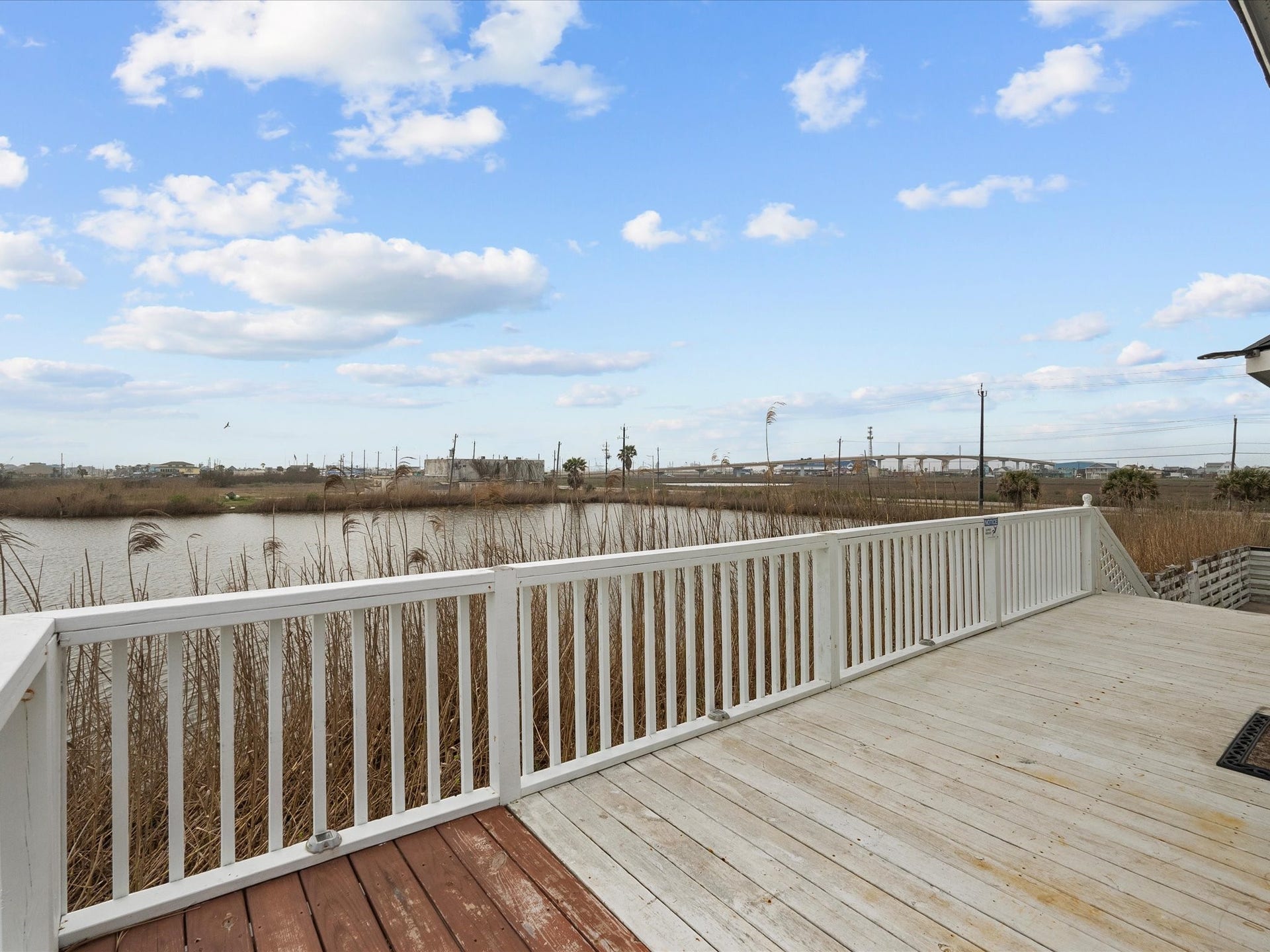 319 Francis Cove, Surfside Beach, TX 77541 Spacious deck with a white railing overlooking a serene waterfront view, featuring reeds and distant structures under a bright blue sky with clouds. Perfect for relaxation and enjoying nature.