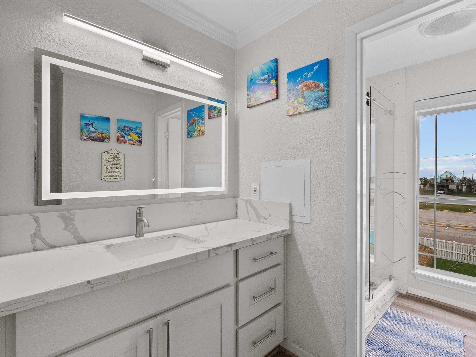 319 Francis Cove, Surfside Beach, TX 77541 This modern bathroom features a sleek marble countertop with an integrated sink, a large backlit mirror, and contemporary fixtures. The room is brightened by natural light from a window with a view, complemented by elegant wall art.
