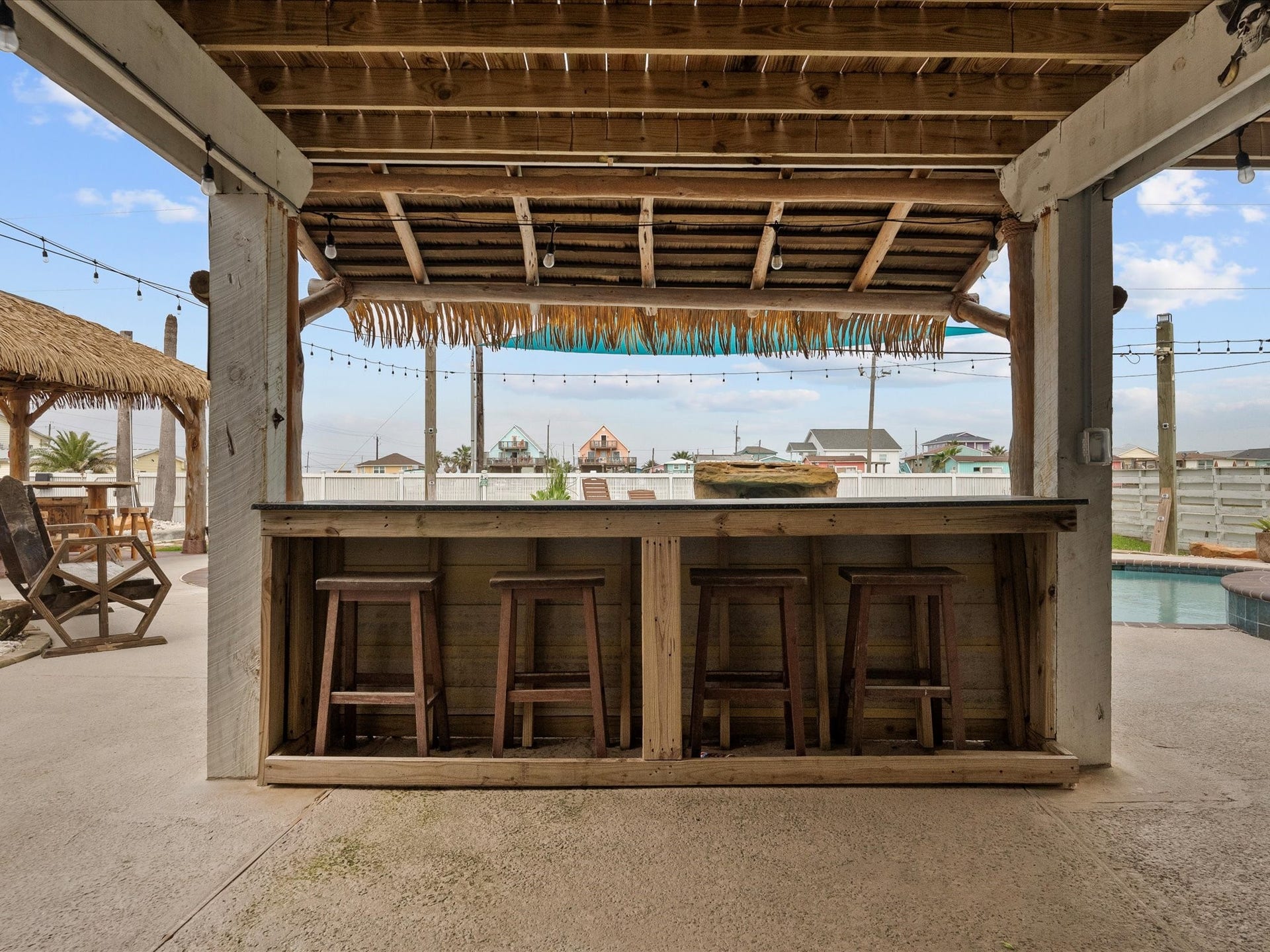 319 Francis Cove, Surfside Beach, TX 77541 This photo showcases an inviting outdoor bar area with wooden stools, a thatched roof, and overhead string lights. It overlooks a pool, creating a perfect space for entertaining and relaxation.