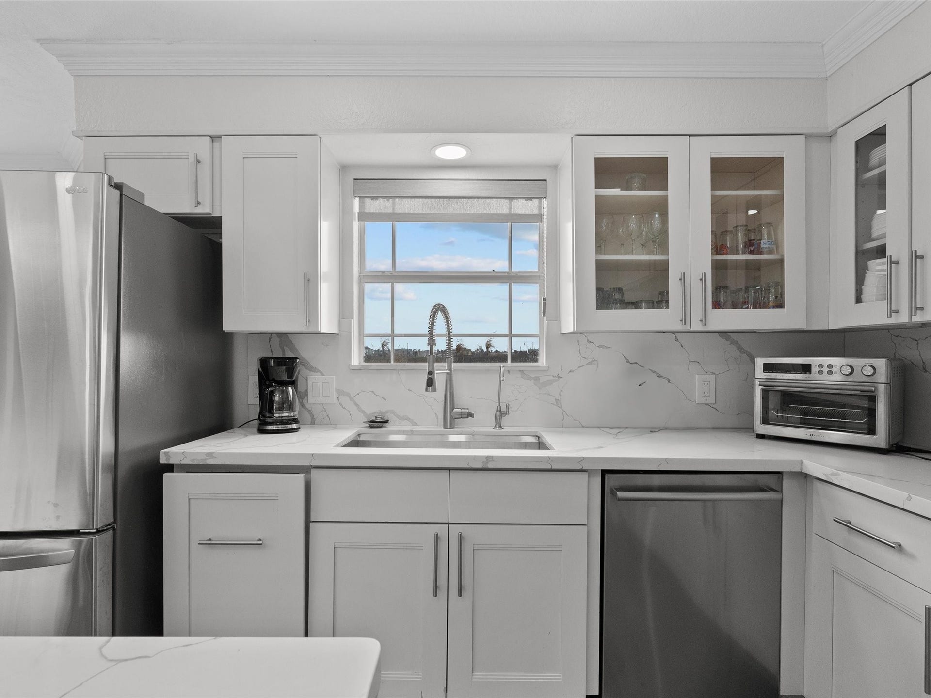 319 Francis Cove, Surfside Beach, TX 77541 This modern kitchen features sleek stainless steel appliances, white cabinetry, and a marble-patterned countertop. It includes a large window above the sink, providing ample natural light, and glass-front cabinets for stylish storage.
