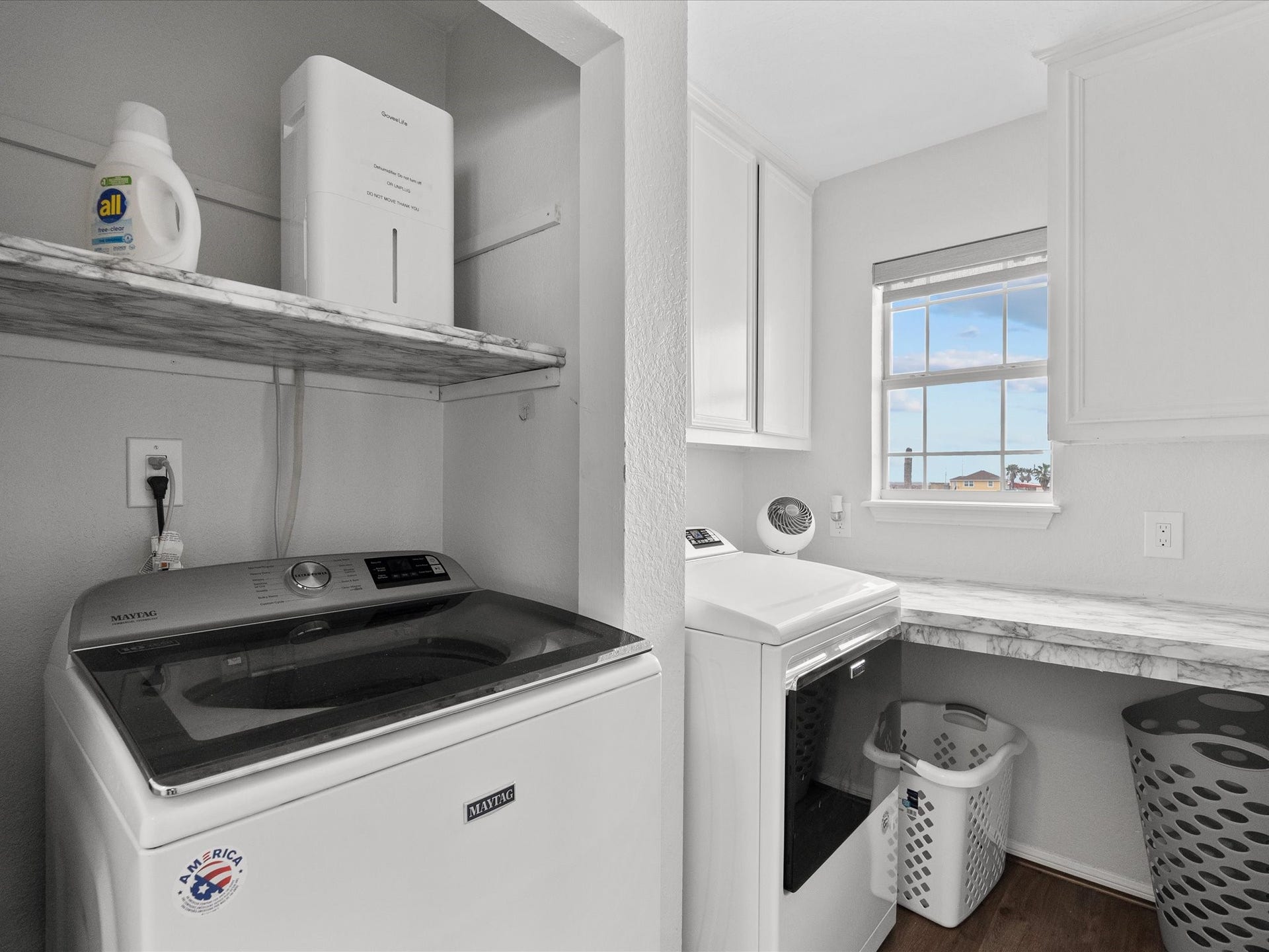 319 Francis Cove, Surfside Beach, TX 77541 This photo shows a bright laundry room with a window, featuring a washer and dryer set. It includes a marble-patterned shelf and countertop, providing ample workspace and storage.