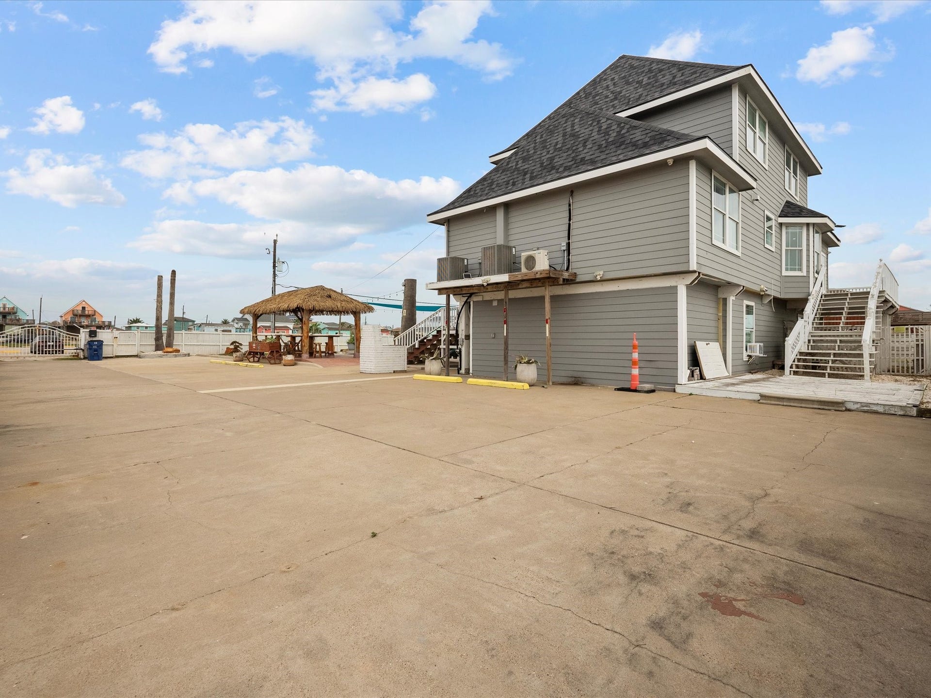 319 Francis Cove, Surfside Beach, TX 77541 This photo shows a spacious waterfront property with a multi-story gray house featuring a sloped roof and exterior stairs. It includes a large paved area and a thatched-roof patio, perfect for outdoor leisure and entertaining.