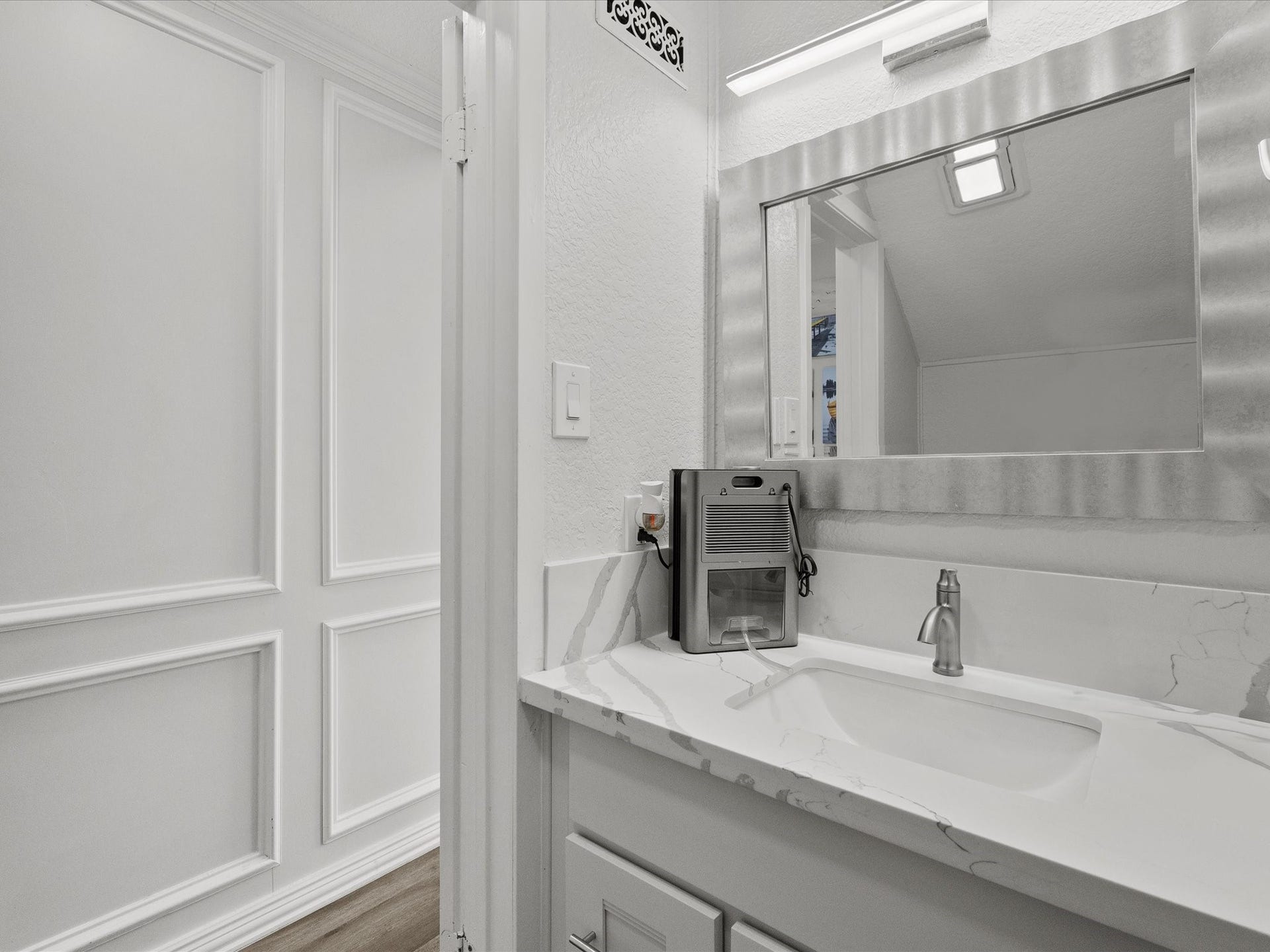 319 Francis Cove, Surfside Beach, TX 77541 This photo showcases a modern bathroom with a sleek white vanity, marble countertop, and elegant paneling. The large mirror and contemporary lighting enhance the space's clean and bright aesthetic.