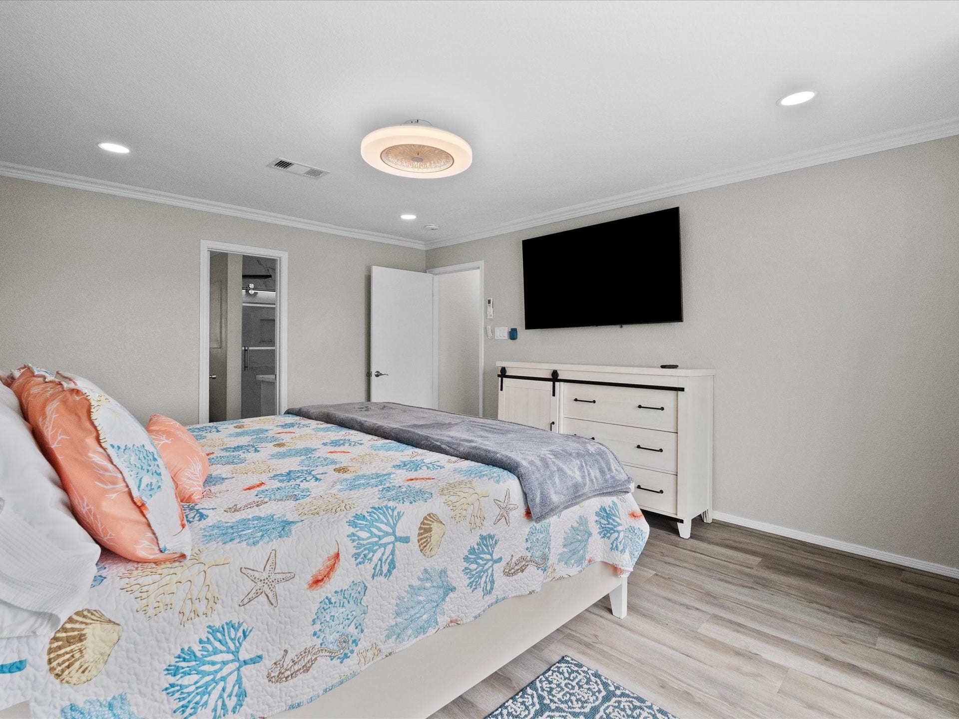 319 Francis Cove, Surfside Beach, TX 77541 Bright bedroom with light wood flooring, a modern ceiling light, and recessed lighting. Features a cozy bed with a colorful sea-themed quilt, a wall-mounted TV, and a dresser with sliding barn door accents. Neutral walls create a calming atmosphere.