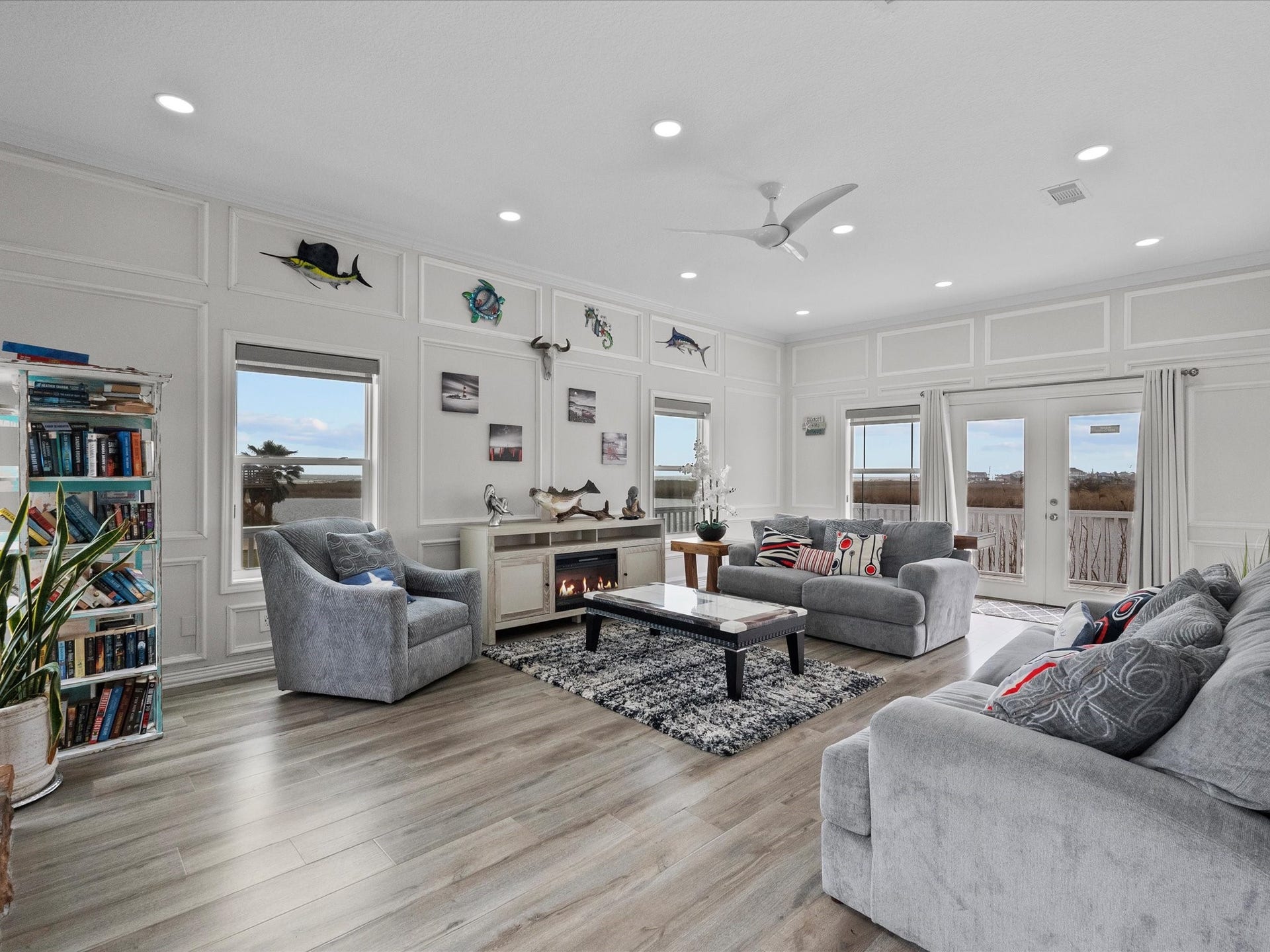 319 Francis Cove, Surfside Beach, TX 77541 This spacious living room features elegant wood flooring, abundant natural light, and a cozy fireplace. Decorated with tasteful accents and ocean-themed art, it includes plush seating and a large rug. French doors open to a scenic outdoor view, enhancing the airy and inviting atmosphere.