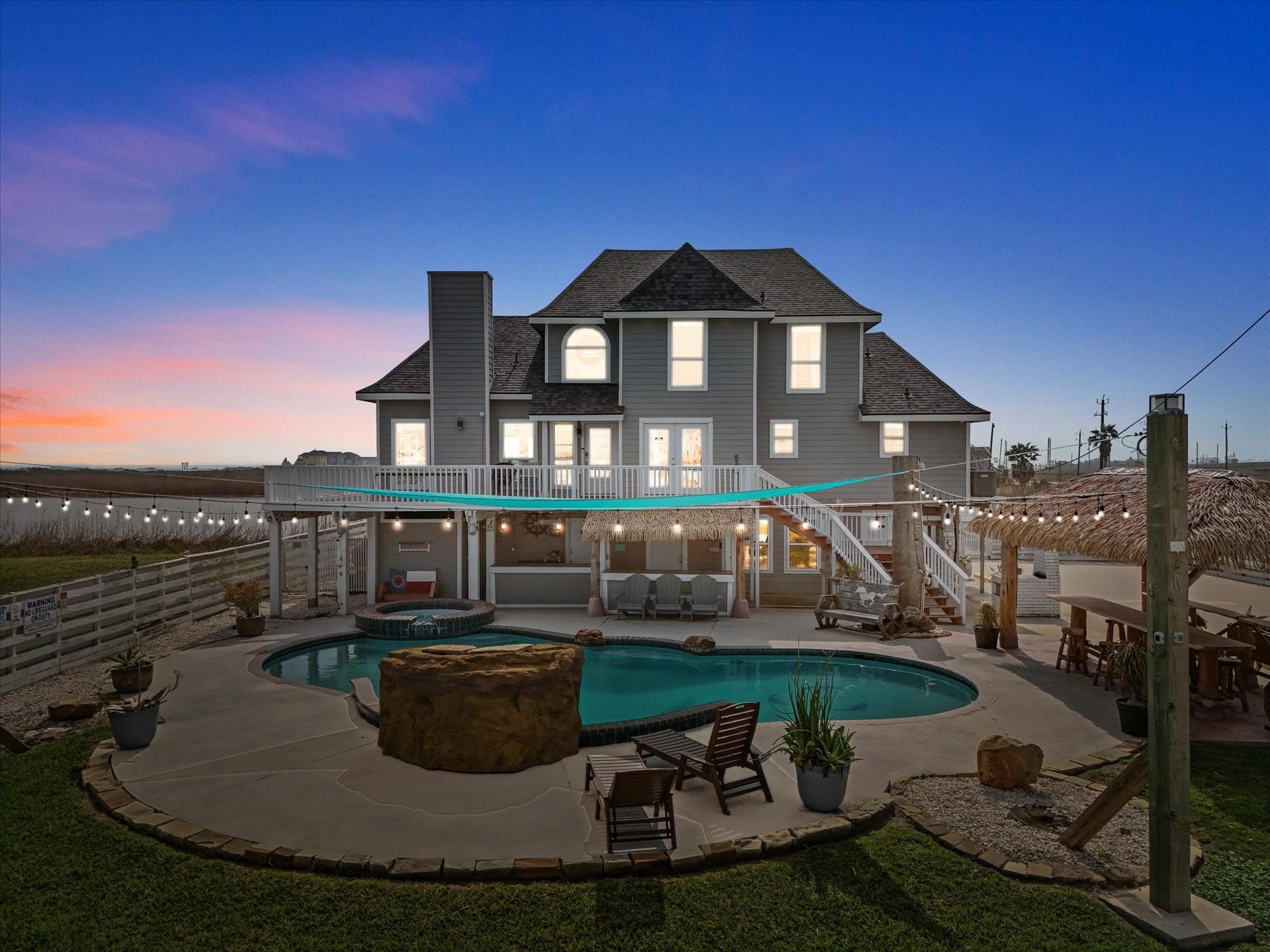 319 Francis Cove, Surfside Beach, TX 77541 This stunning home features a spacious backyard with a beautiful pool and hot tub, surrounded by elegant landscaping. The multi-story house includes a large deck, perfect for entertaining, and is illuminated against a picturesque evening sky.