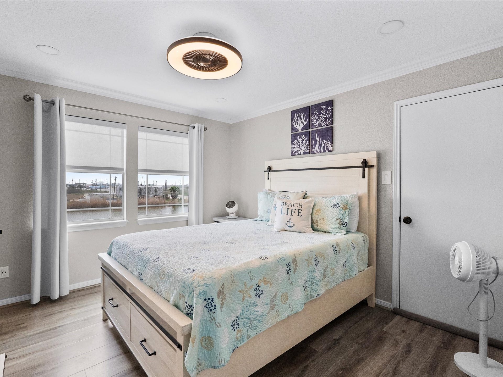 319 Francis Cove, Surfside Beach, TX 77541 This cozy bedroom features a modern ceiling light, large windows with views, and a beach-themed decor. The room includes a comfortable bed with storage drawers and light wood flooring, creating a bright and inviting space.