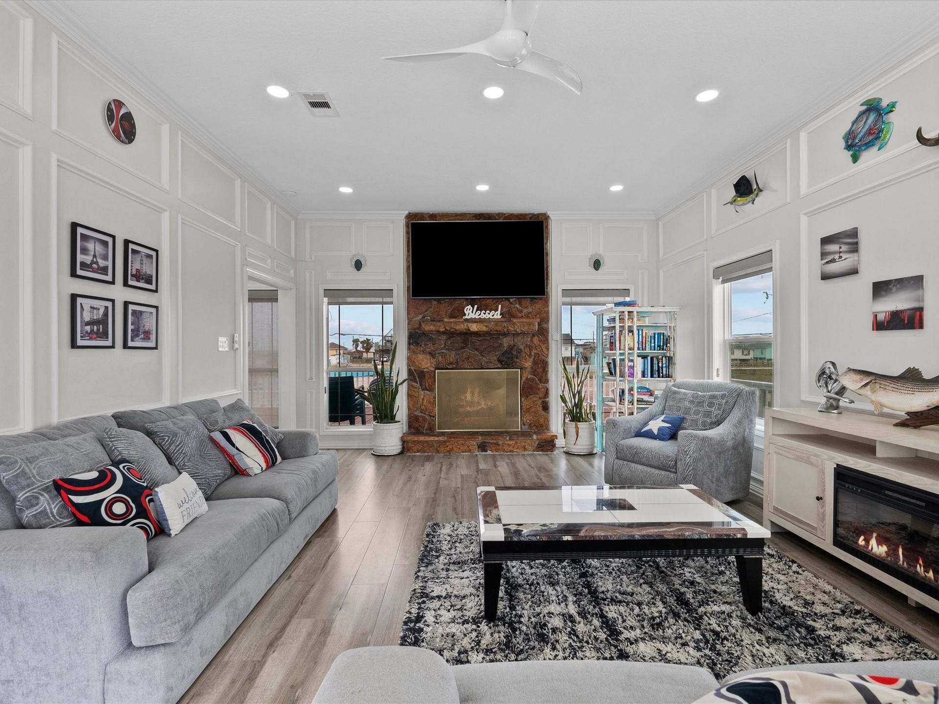 319 Francis Cove, Surfside Beach, TX 77541 This living room features a cozy and stylish design with light gray sofas and a patterned rug. A stone fireplace with a mounted TV serves as the focal point, while abundant natural light streams in through large windows. The room is accented with modern decor and a ceiling fan, creating a welcoming atmosphere.