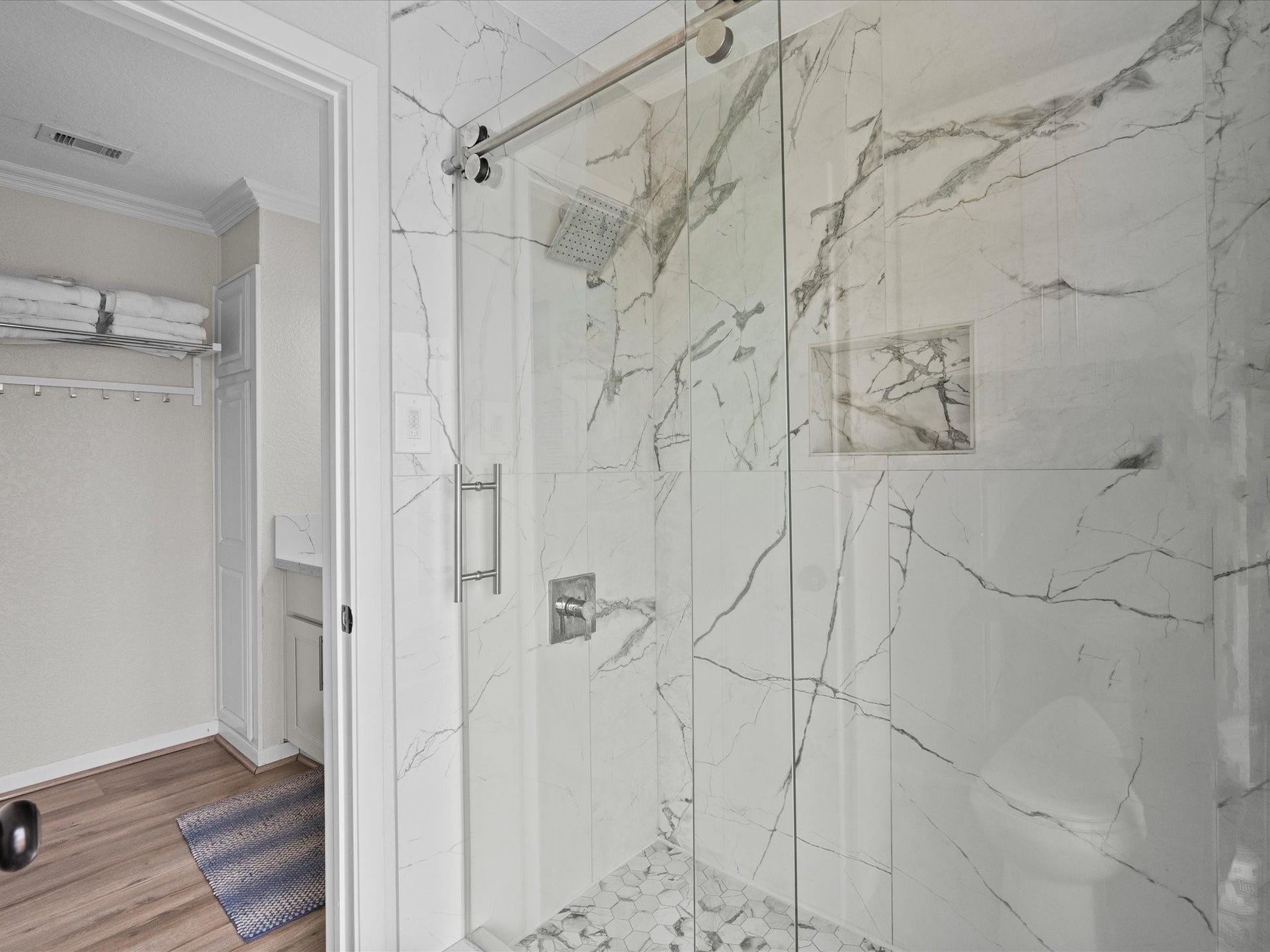 319 Francis Cove, Surfside Beach, TX 77541 Modern bathroom with a sleek glass-enclosed shower featuring elegant marble walls and a pebble floor. Adjacent space includes storage shelves and a vanity area, accented with light wood flooring.