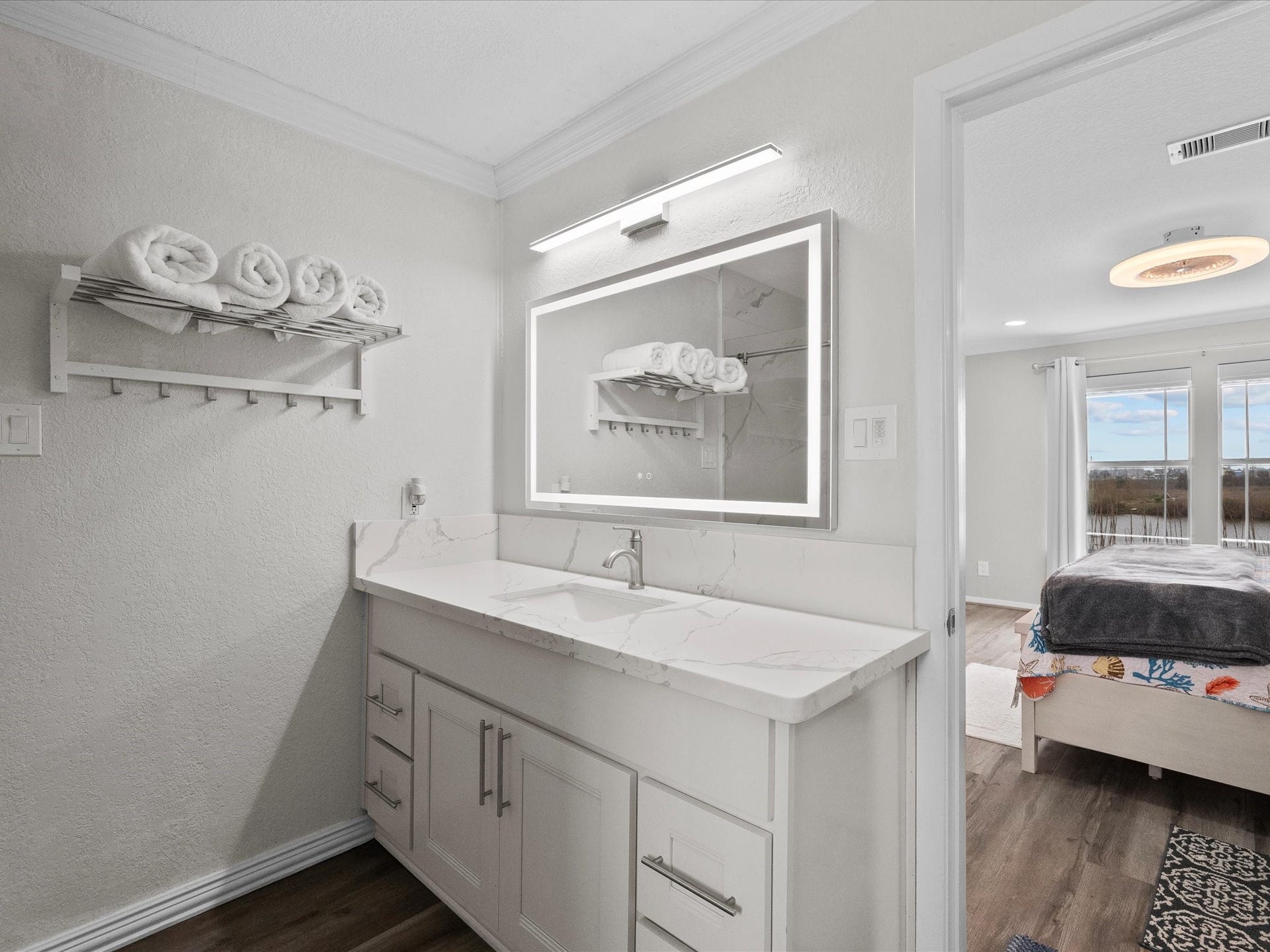 319 Francis Cove, Surfside Beach, TX 77541 Modern bathroom with a sleek vanity and illuminated mirror, featuring a towel rack with neatly rolled towels. It opens into a bright bedroom with large windows and a scenic view.