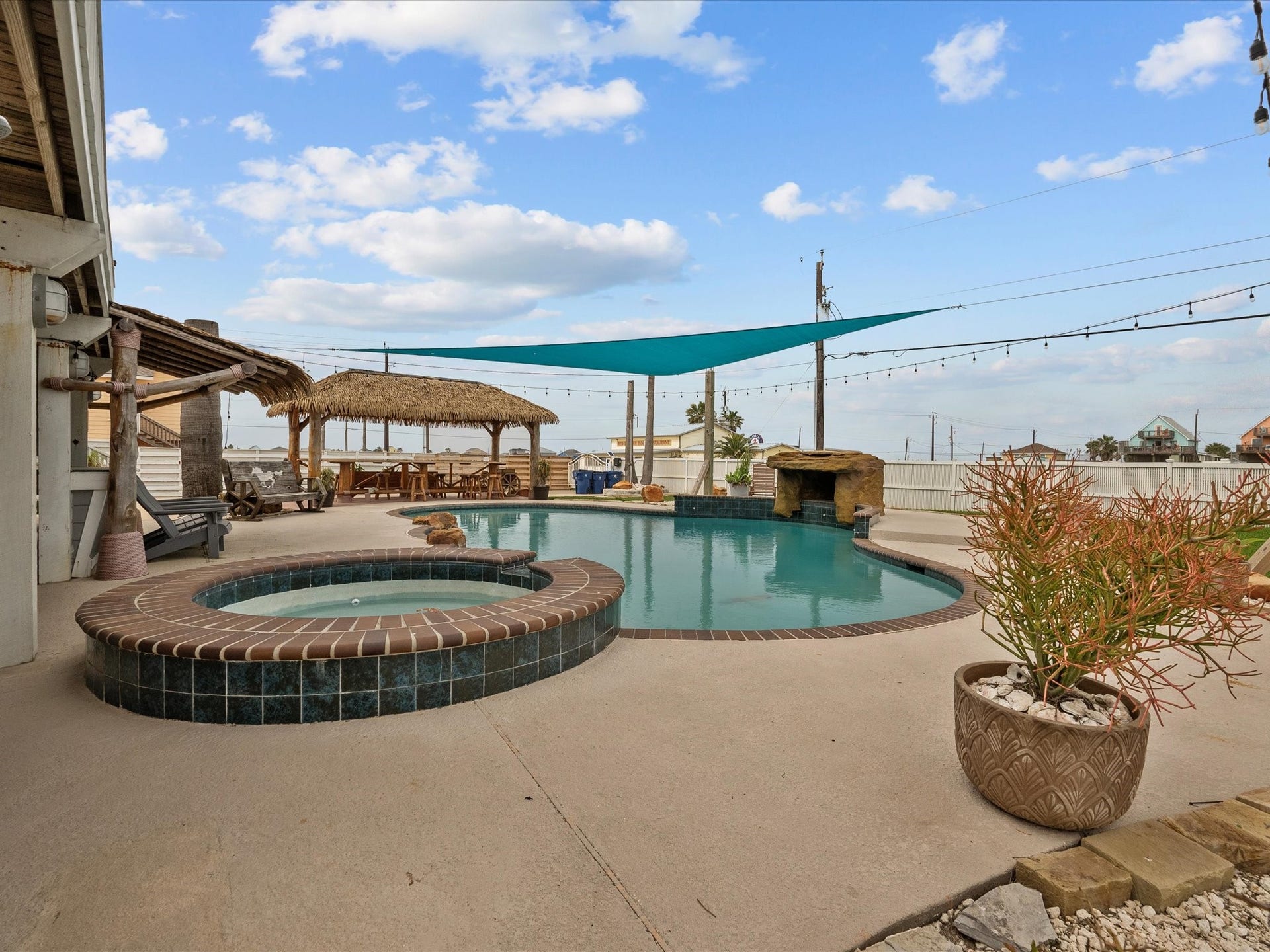 319 Francis Cove, Surfside Beach, TX 77541 This backyard oasis features a spacious pool with a connected hot tub, shaded by a sail canopy. There's a charming tiki-style hut for outdoor lounging, surrounded by string lights, offering a perfect space for relaxation and entertaining.
