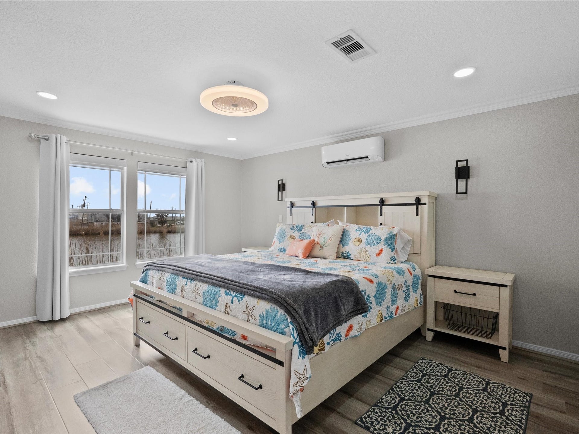 319 Francis Cove, Surfside Beach, TX 77541 This bright and airy bedroom features a large bed with coastal-themed bedding, light wood flooring, and neutral walls. There's ample natural light from two windows, modern recessed lighting, a stylish ceiling fixture, and a wall-mounted air conditioning unit for comfort.