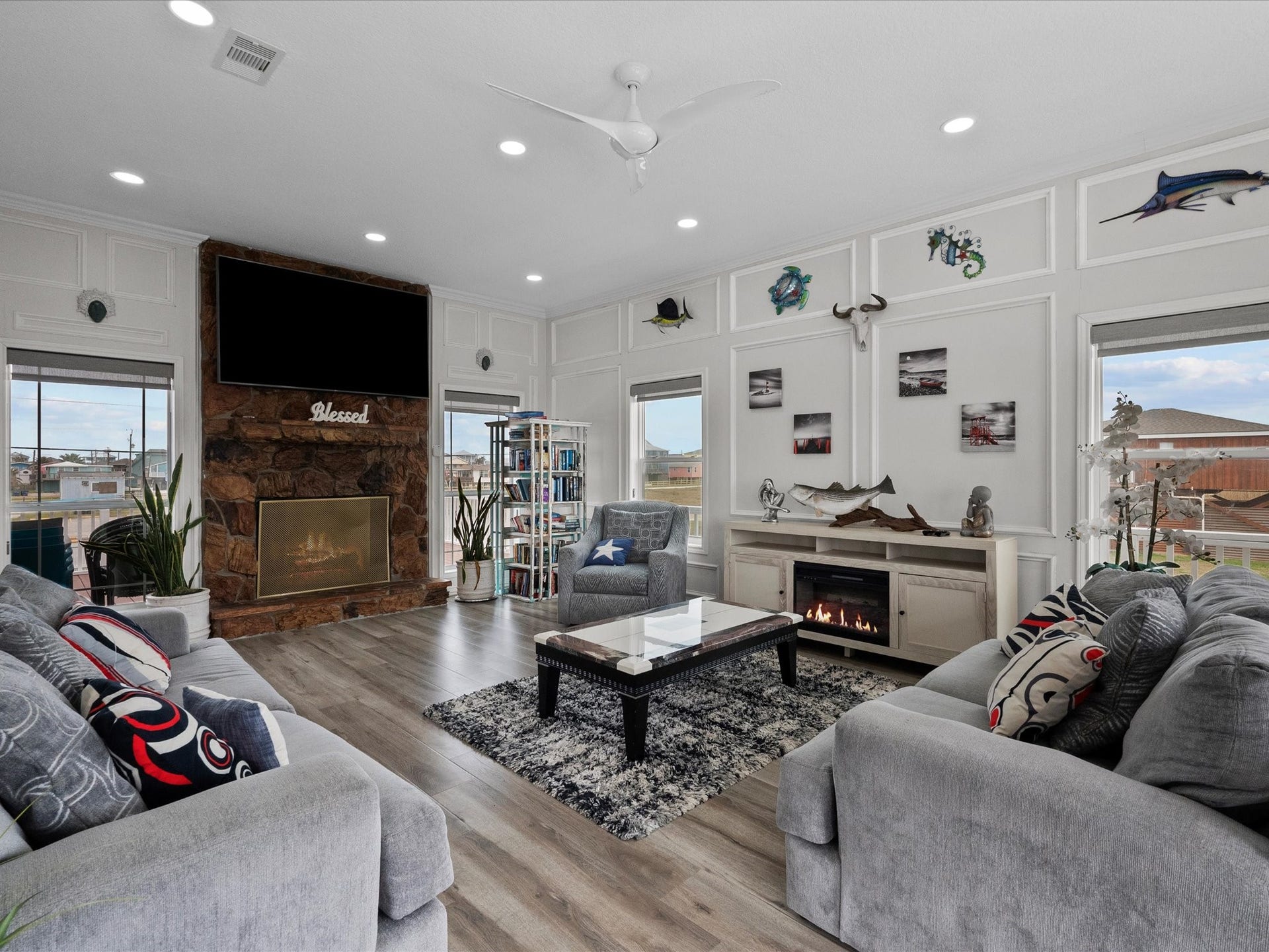 319 Francis Cove, Surfside Beach, TX 77541 This living room features modern decor with a cozy fireplace, large windows for natural light, and stylish wood flooring. It includes a stone accent wall with a mounted TV, comfortable gray seating, and nautical-themed decor, creating a welcoming and relaxing atmosphere.