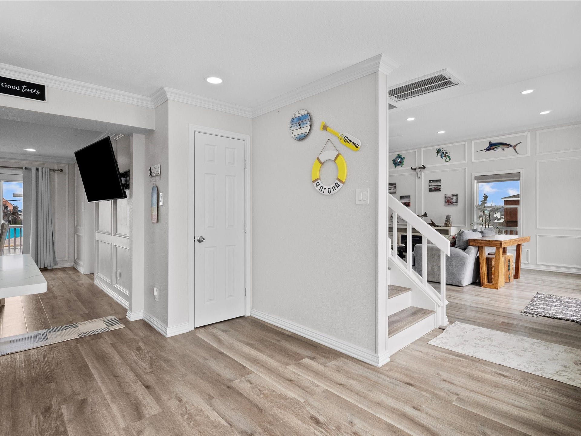 319 Francis Cove, Surfside Beach, TX 77541 This photo showcases a bright, open-concept living area with modern wood flooring and neutral walls. The space features a staircase, wall-mounted TV, and decorative wall accents. Large windows provide ample natural light, and there's a view of the outdoors.