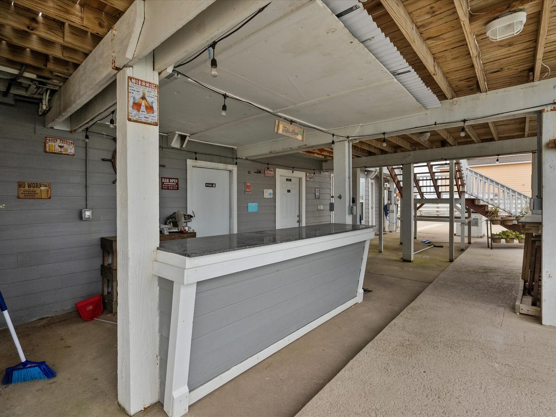 319 Francis Cove, Surfside Beach, TX 77541 This photo shows an outdoor bar area under a wooden deck, featuring a countertop with overhead string lighting. There's adjacent stair access and decorative signs, creating a casual, inviting space perfect for entertaining.